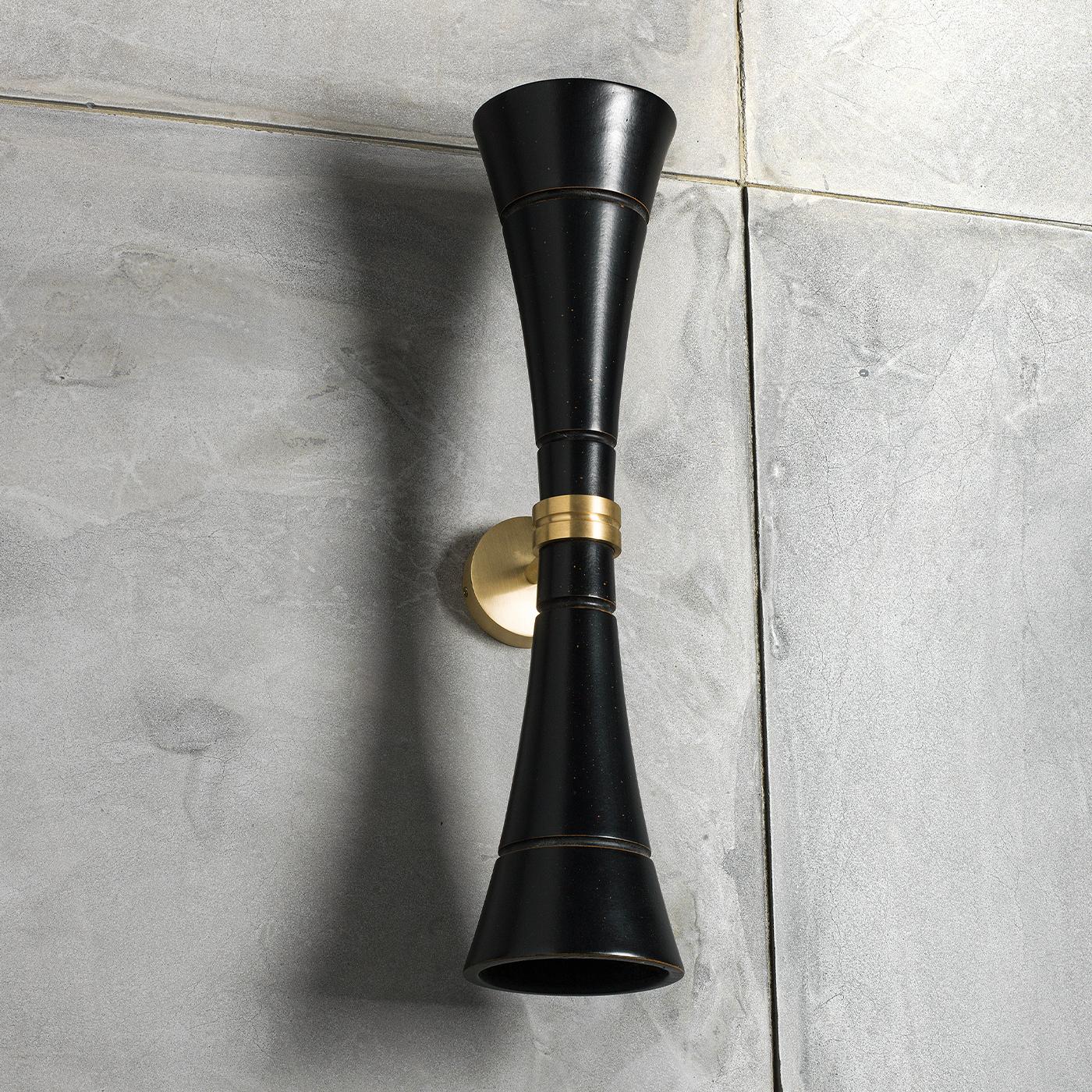 A unique wall lamp characterized by the double cone-shaped element that houses a LED light at both of its ends. This is to project the light both upwards and downwards. The element is made in antiqued-black painted wood and it is supported by a