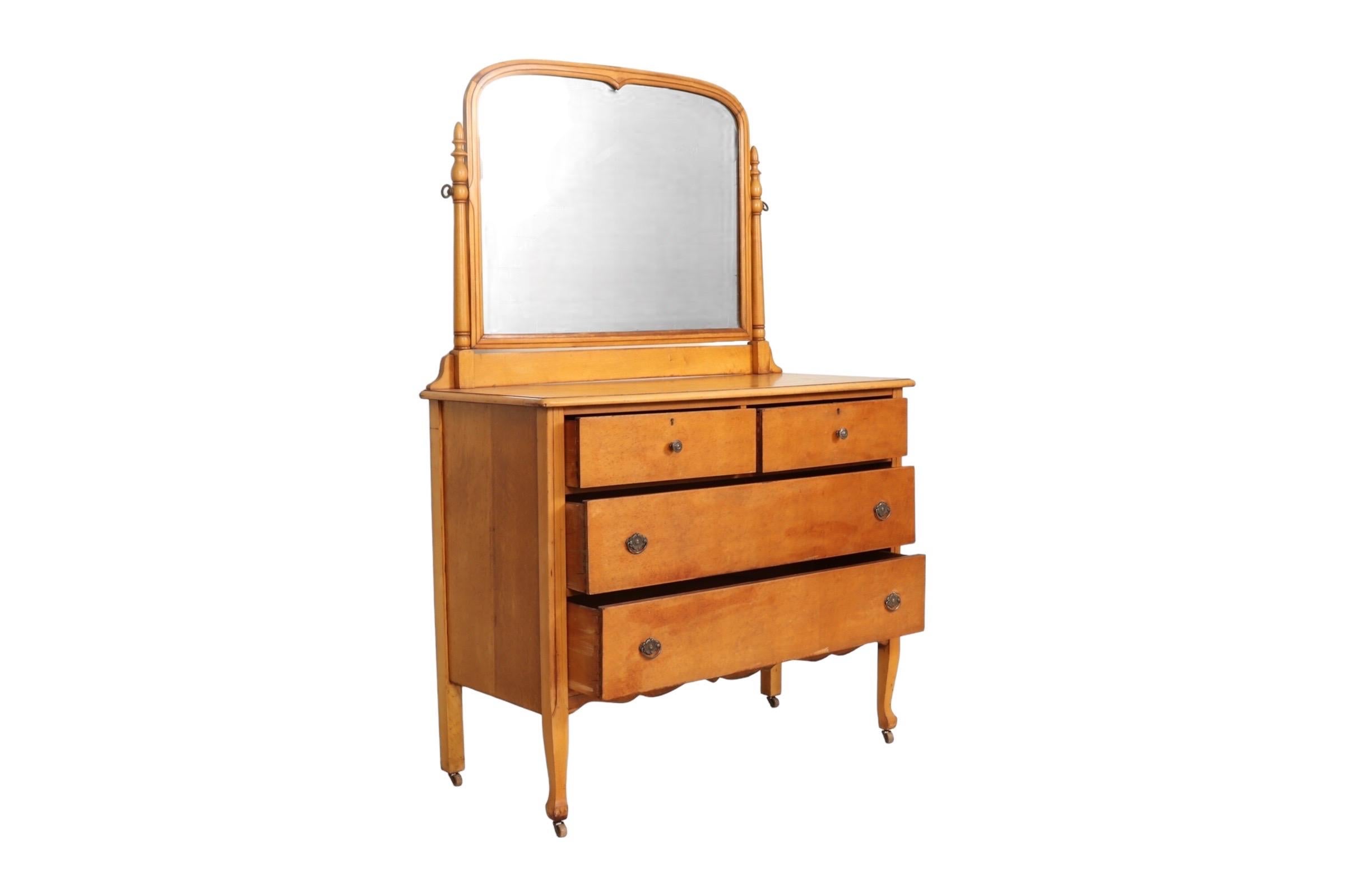An antique two over two vanity dresser in birdseye maple made by Conewango Furniture Co of Warren, Pennsylvania. An arched mirror set in a simple beveled frame is supported with acorn topped turned posts. Dovetailed drawers open with simple cast