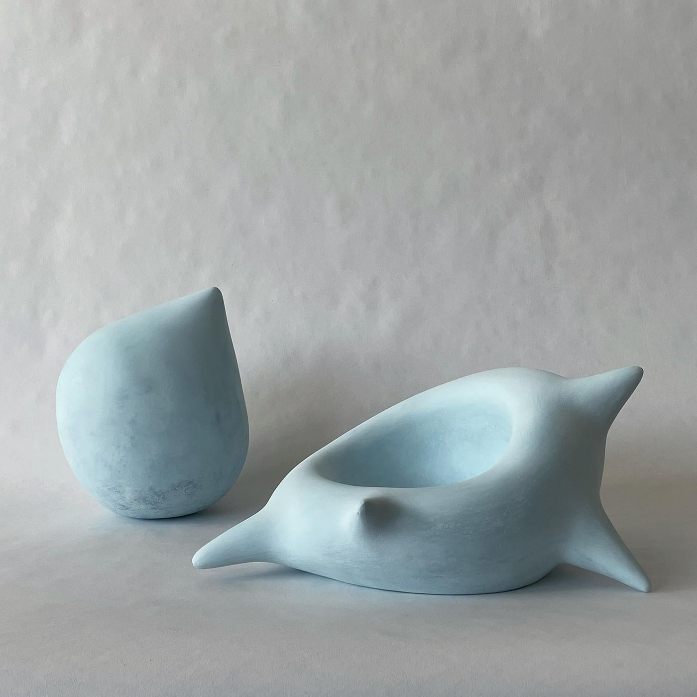 La scultura in ceramica azzurra Confidente ha una forma semplice e leggermente inclinata con sporgenze arrotondate. La sua patina stratificata di blu e celeste crea un senso di profondità e morbidezza. La scultura appare attenta, favorendo una