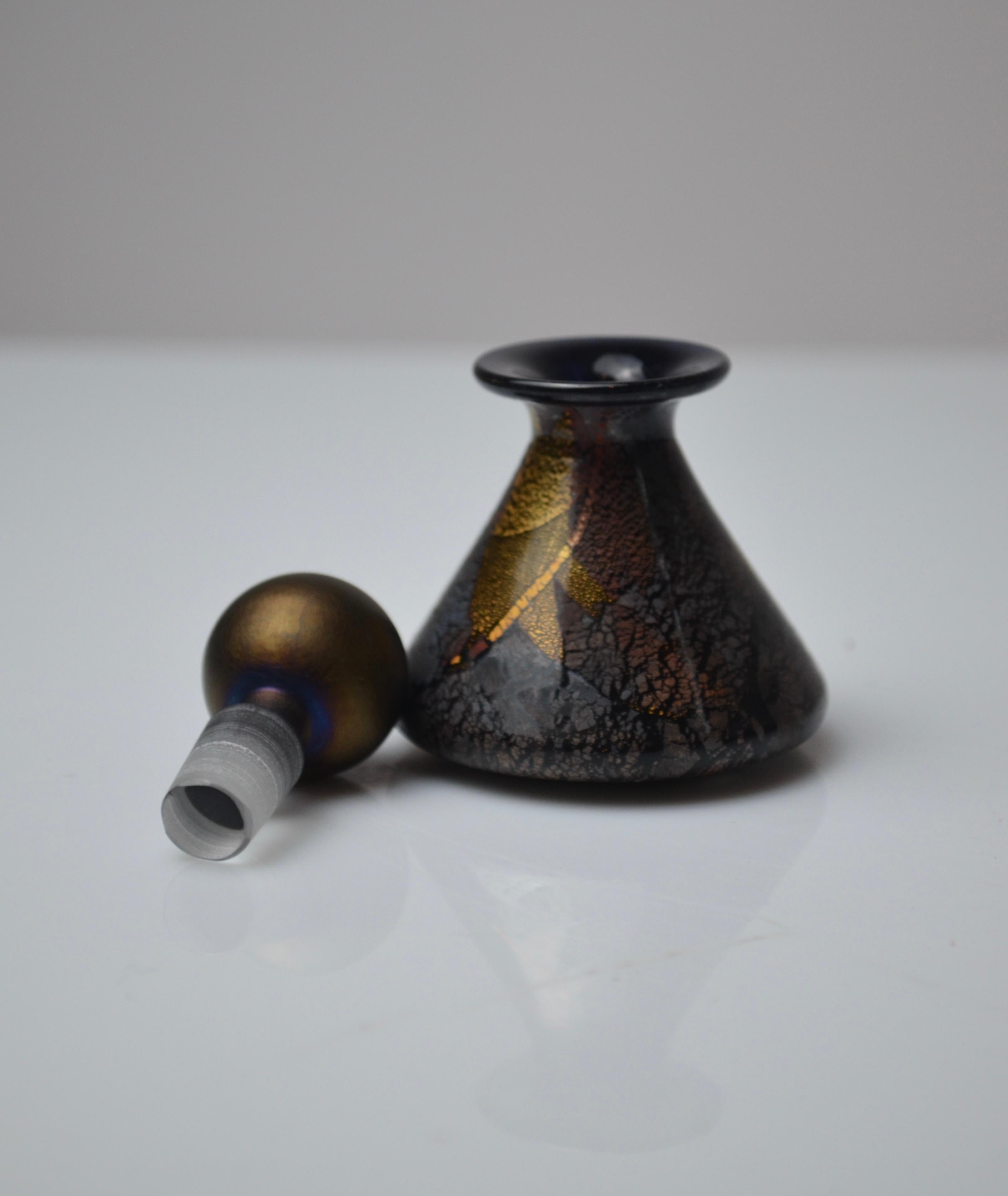 Elegante frasco de perfume cónico pequeño de vidrio soplado, fabricado por el renombrado Studio A de la Isle of Wight, fundado por Michael Harris.
Esta pieza presenta una llamativa superficie craquelada en tonos oscuros, resaltada con brillantes