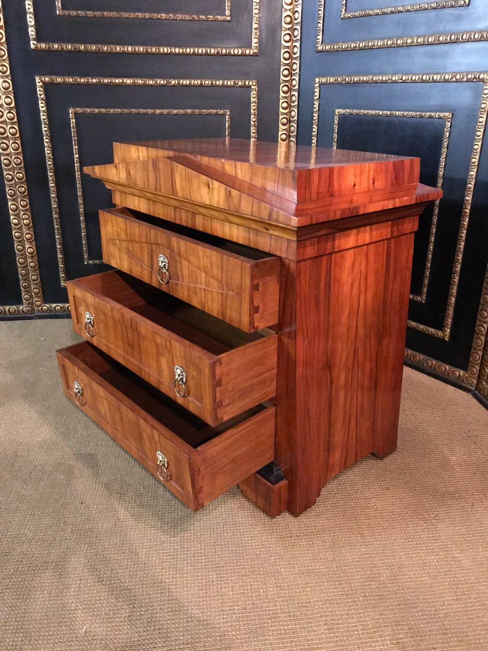 Mahogany on solid conifers. Conical body on profiled base. In the front, three casted drawers. Overhanging grooved profile inserts flat gusset, stepped with oval cover plate. Solenoid head fittings with grip rings. So called pyramid furniture are