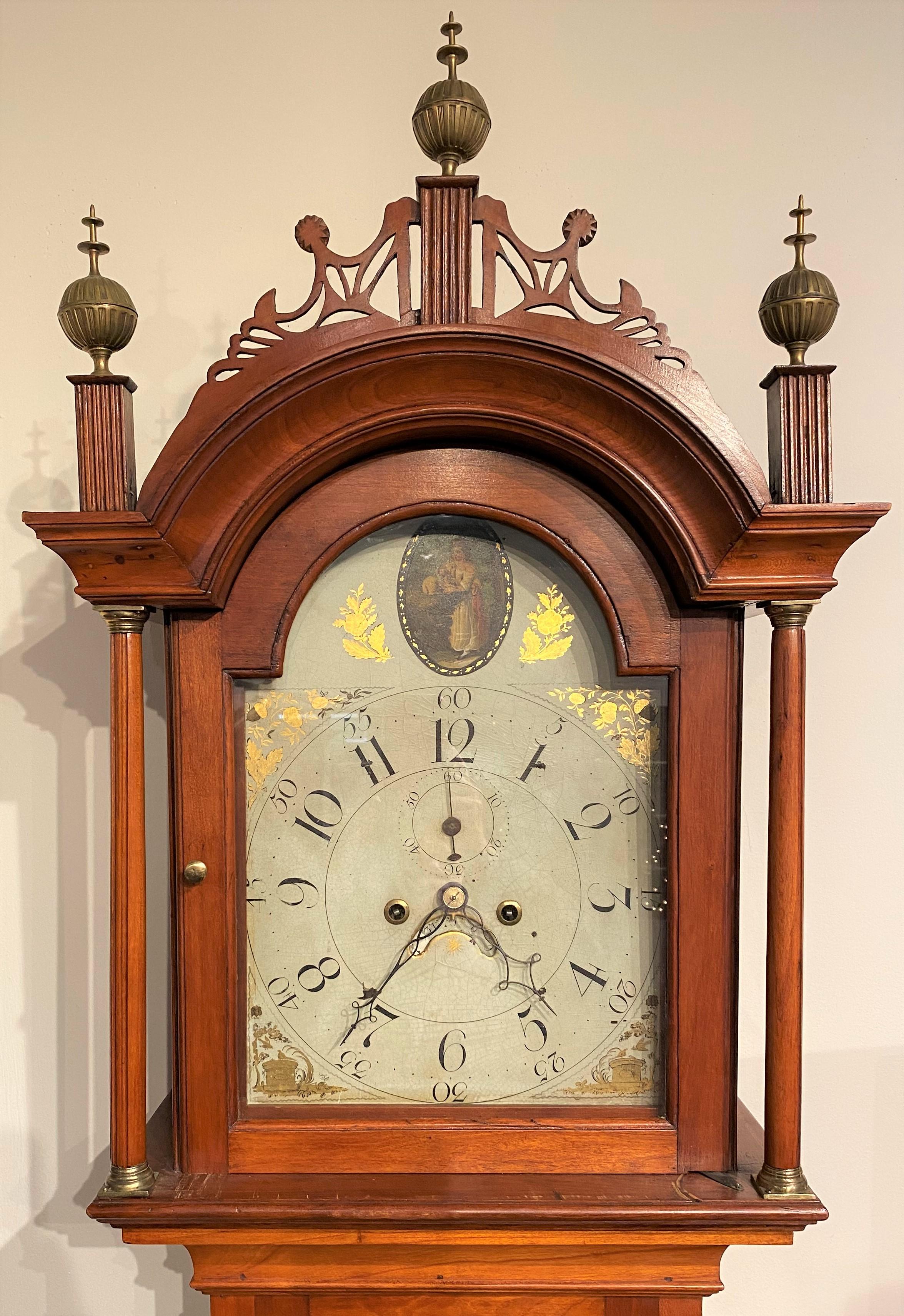 A fine cherry wood tall case clock with fretwork carved crest on the bonnet, along with three spire and ball finials with reeded wooden plinths, surmounting a glass arched door, opening to a metal faced hand painted arched dial with an oval image of