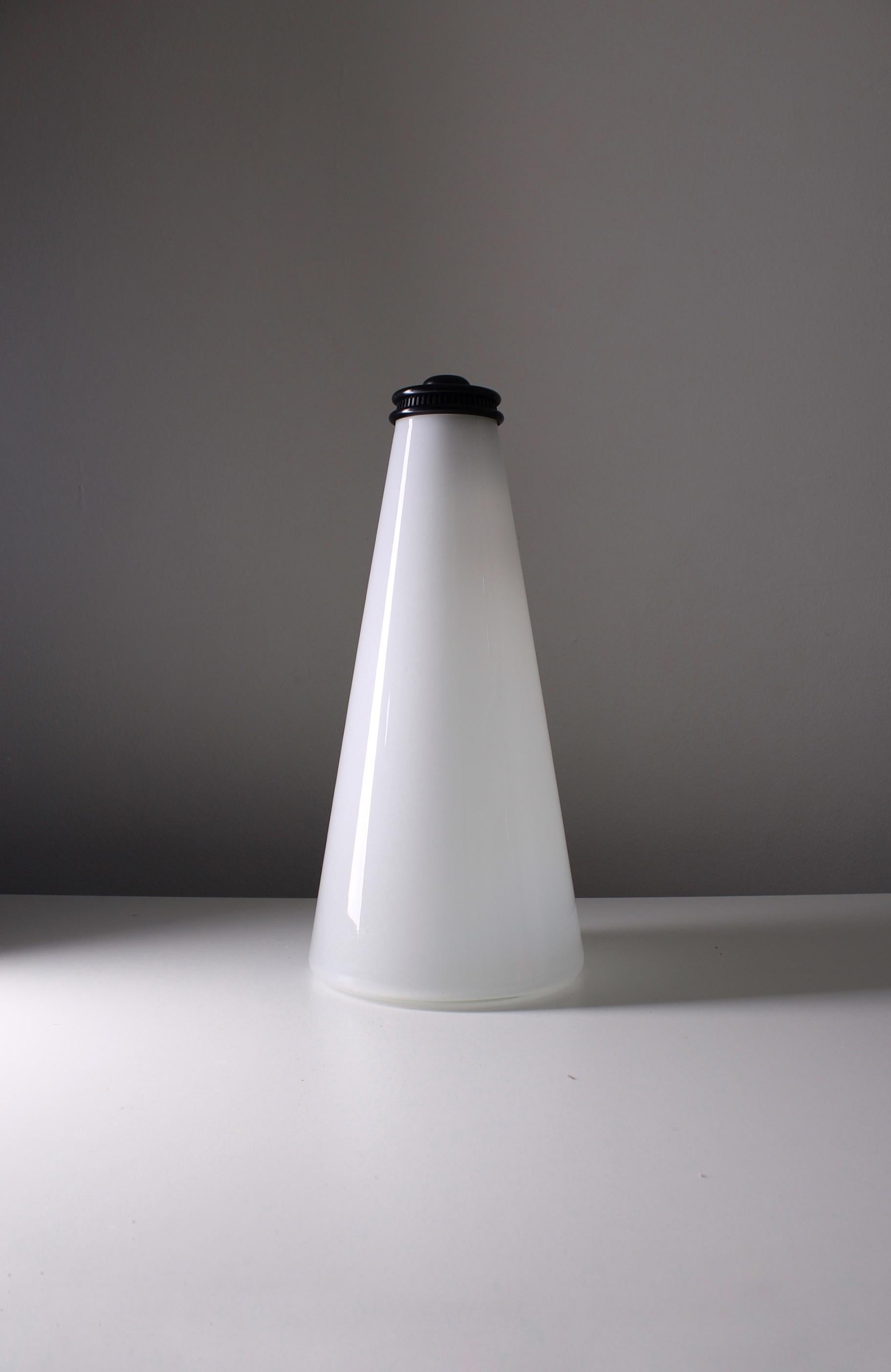 
Table lamp, model Cono designed by Ezio Didone for Arteluce in 1979. The lamp is made of opaque milky-white Murano glass with a subtle gradient. On the top of the lamp is a round black cap that holds the armature and a switch. The lamp was produced