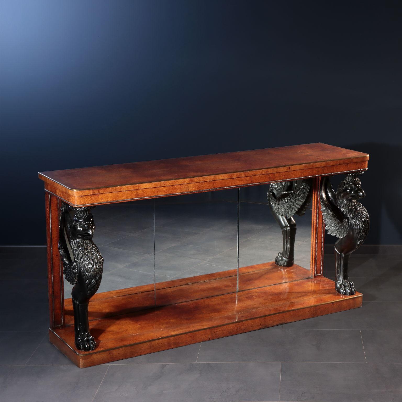 English console resting on a shelf and equipped with two monopod griffins shaped uprights; the rear panel consists of three mirror plates. Entirely veneered in amboina briar, it has a bronze frame and brass covered top edges.