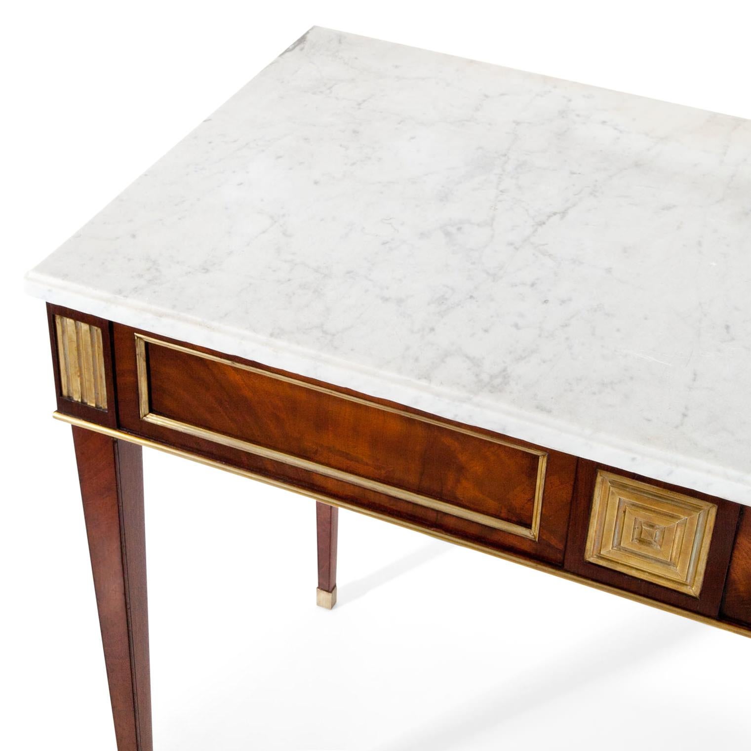 Console table standing on tall tapered legs with brass caps. The straight apron shows fillings with brass molding as well as geometrical applications. The white marble-top is recent.