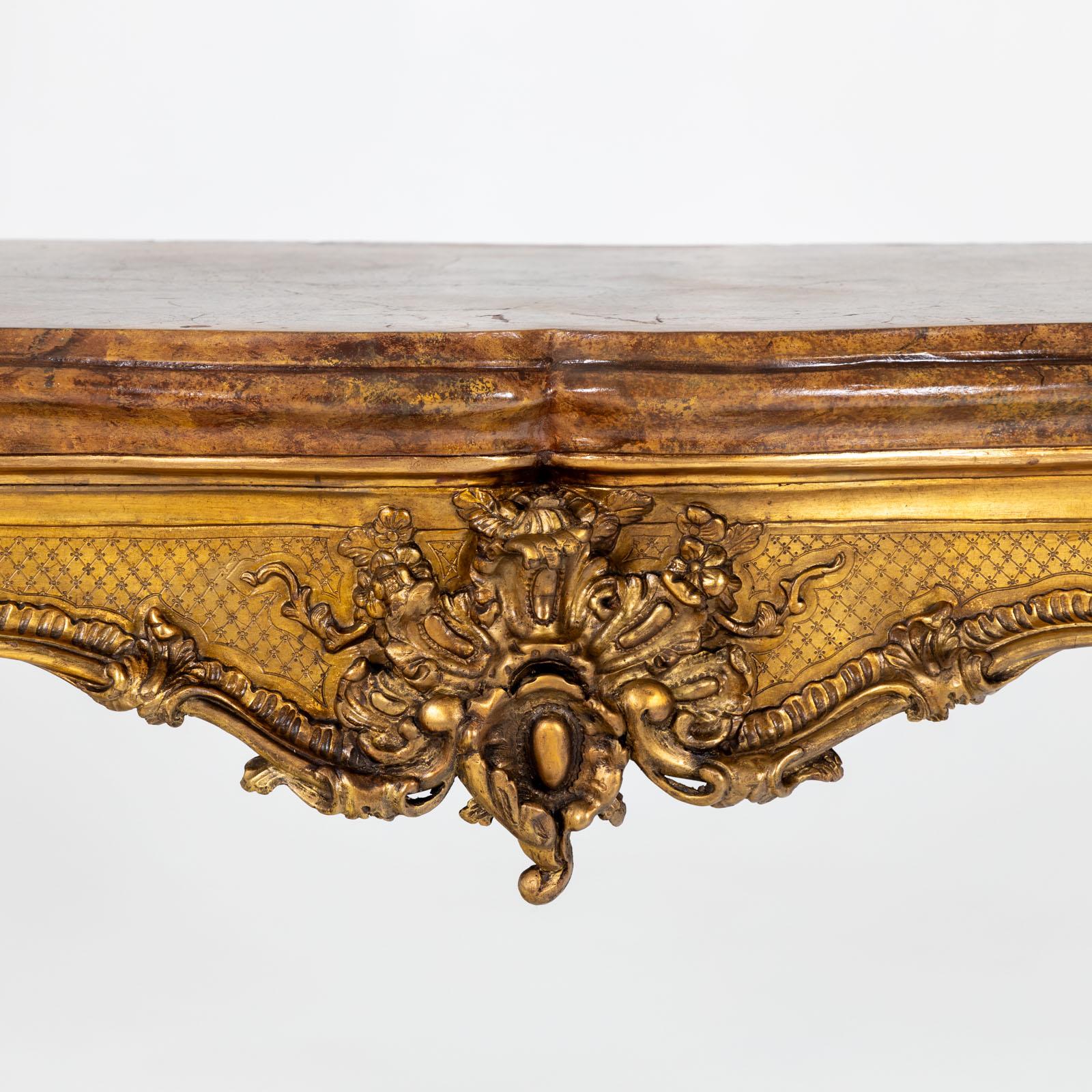Baroque-style wall console with a richly decorated and gold-patinated frame with s-shaped legs and rocailles as well as shell decoration on the curved frame. The ochre-coloured table top in scagliola technique is curved and profiled several times. 