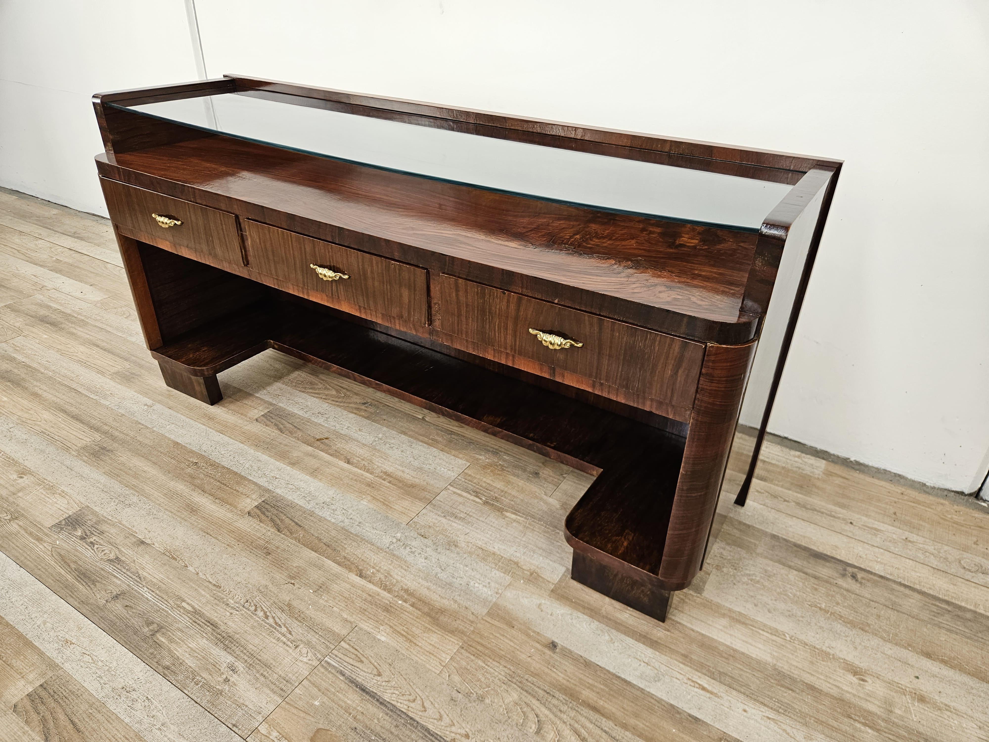 Refinado tocador o consola Art Déco de raíz de nogal de principios de los años 40, un mueble con múltiples usos gracias a sus reducidas dimensiones y su diseño distintivo.

Tiene una tapa de cristal transparente con tres pequeños cajones decorados