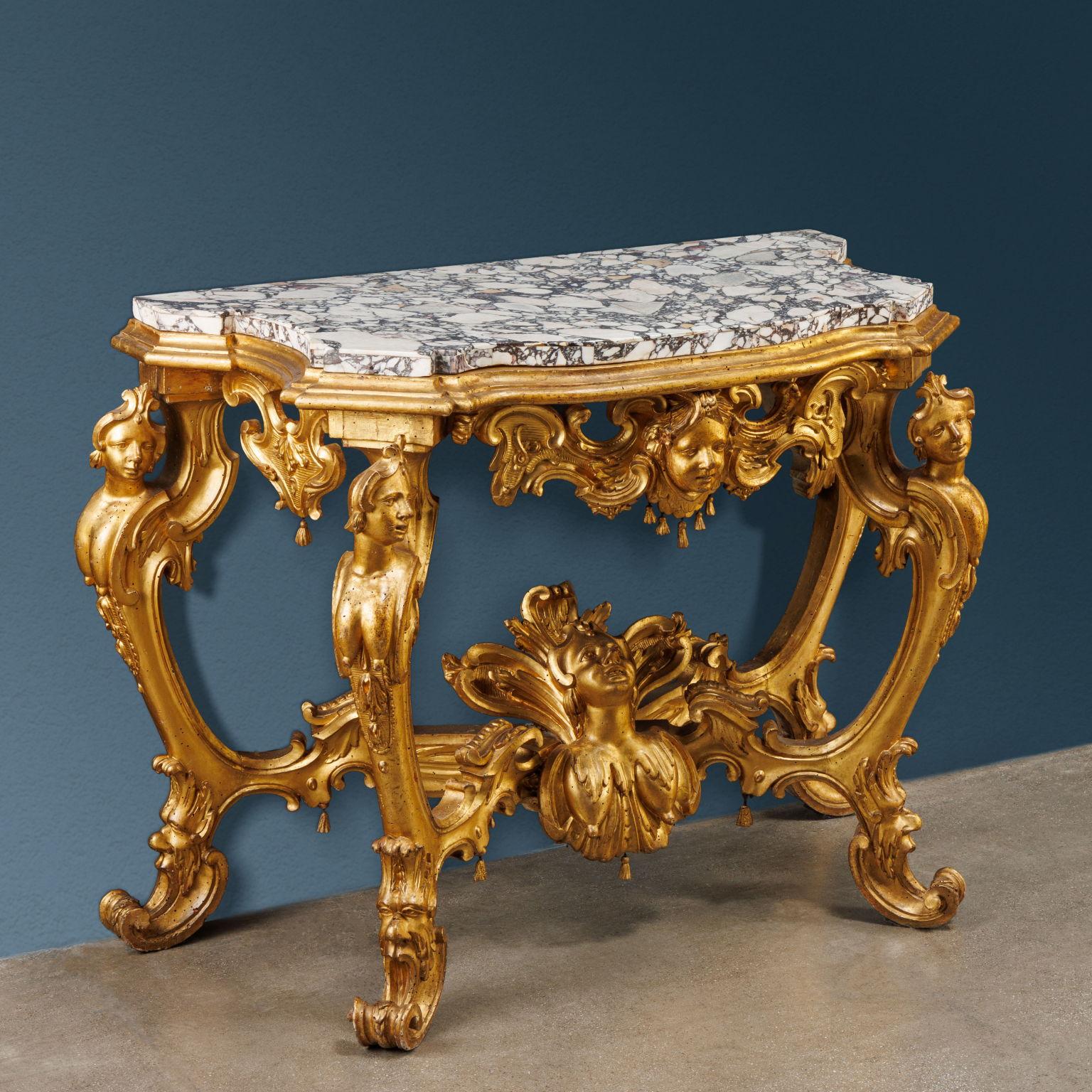 Carved and gilded wooden console table. It rests on four opposing double-curved supports ending in full-bodied curling volutes held by masks; carved female caryatids rest on the upper volutes in the round. The connection of the legs is made by