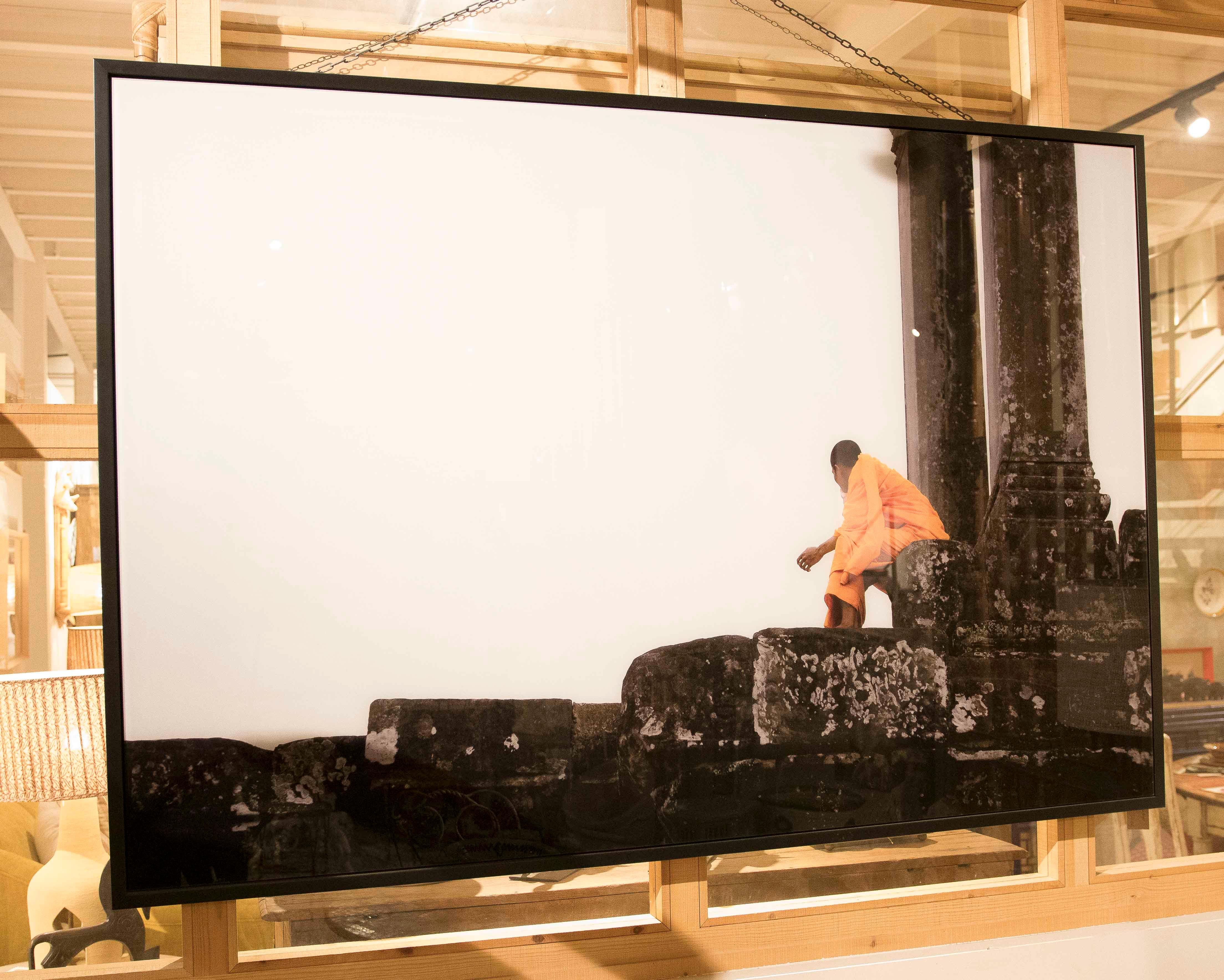 Photographie saisissante d'un moine vêtu d'une robe orange vif, assis en silence sur les pierres usées d'un temple historique. Le contraste entre le minimalisme du fond blanc et la texture des pierres crée une composition visuellement puissante,