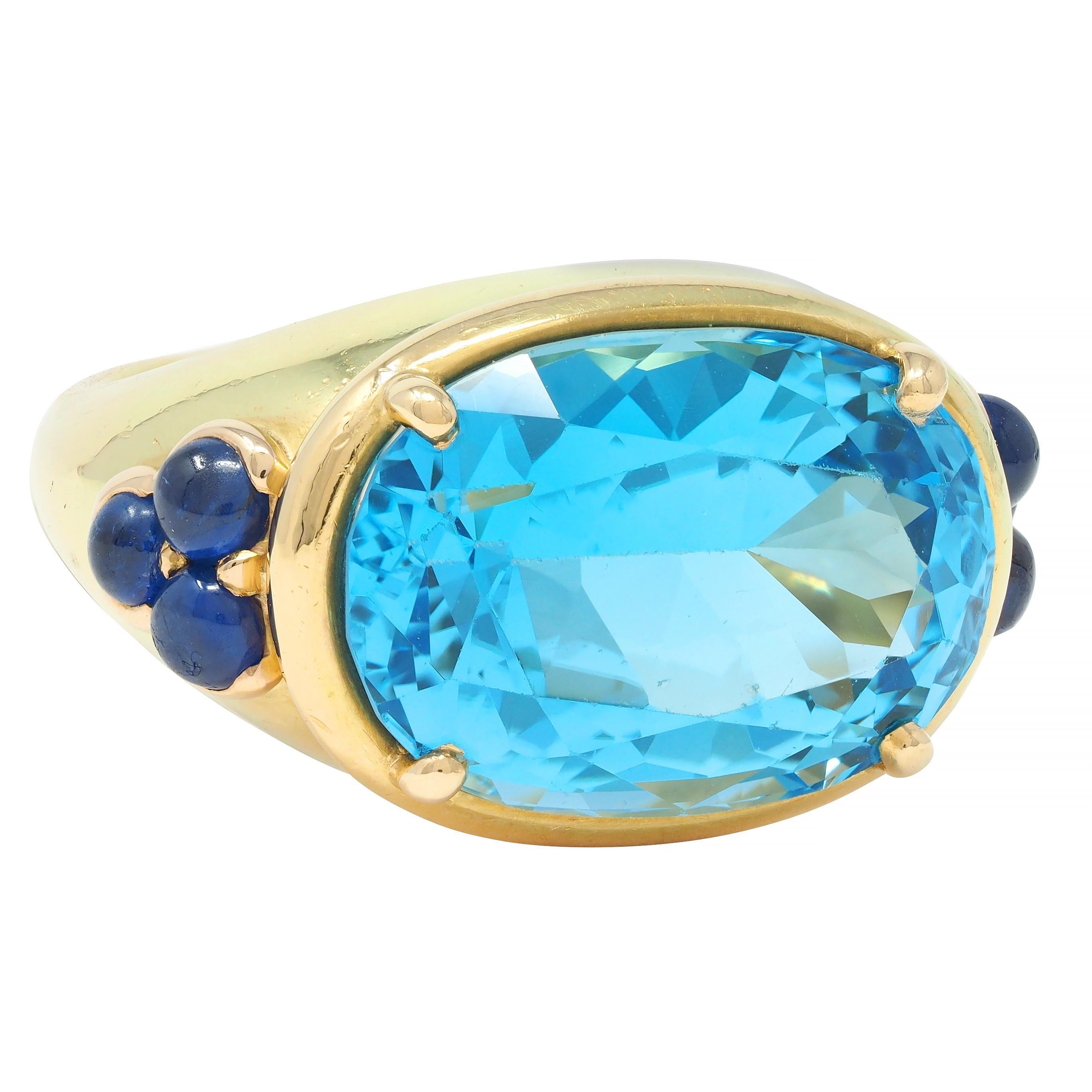 Centering an oval cut topaz weighing approximately 11.01 carats 
Transparent bright medium blue and  prong set east to west
With fluted surround flanked by clusters of bezel set sapphires
Round cabochons weighing approximately 1.04 carats