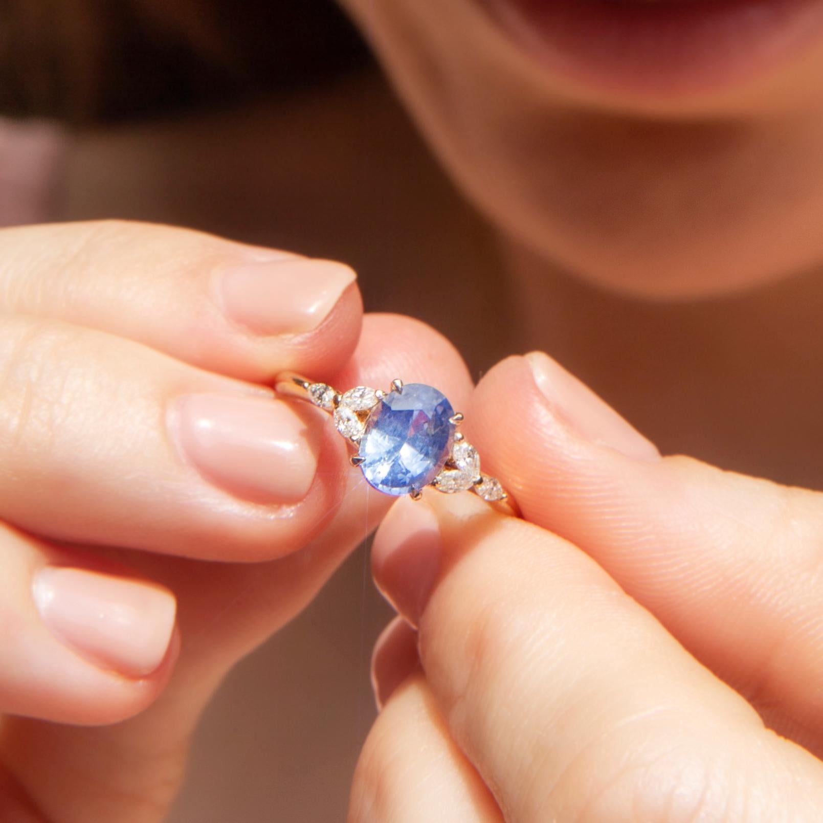 Crafted in 18 carat yellow gold, this adorable contemporary ring features an alluring bright blue Ceylon-type sapphire carefully set between two shoulder-set clusters each holding three marquise cut diamonds glistening in unison. Her name is The