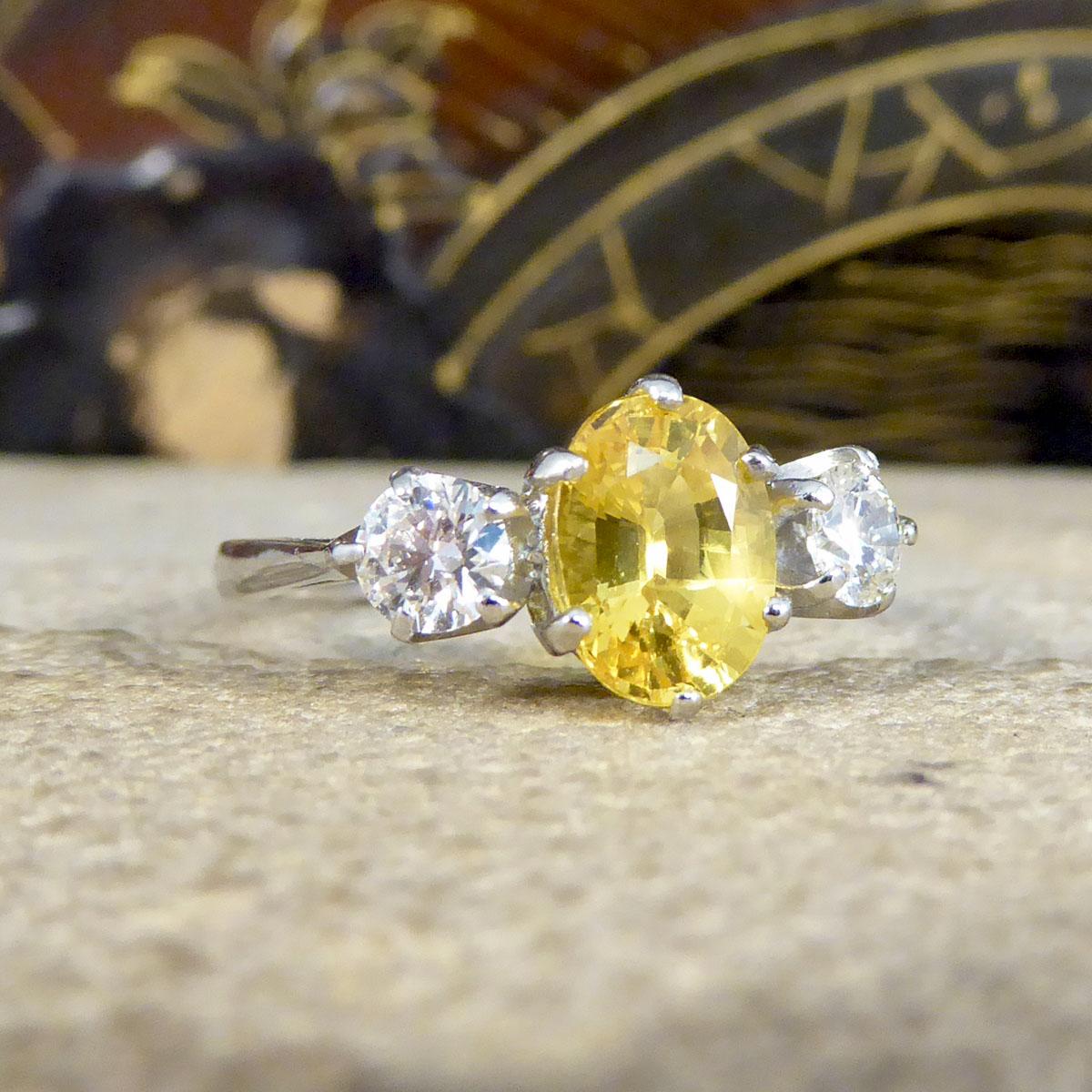 An absolutely gorgeous and sparkly Yellow Sapphire and Diamond three stone ring holds three beautiful stones set in a secure Platinum claw setting. In the centre sits a lovely bright Oval Cut Yellow Sapphire in the centre weighing 1.84ct with a