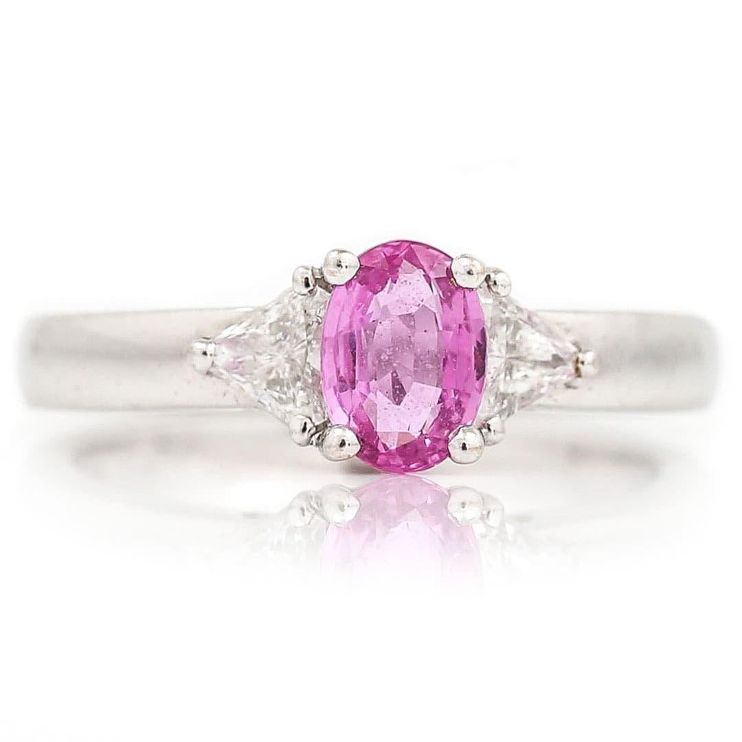A pretty 18ct white gold and central set 0.84ct oval pink sapphire ring which is flanked either side by two trillion cut diamonds totalling approx 0.60ct. The gemstones are claw set and the ring carries a full UK ​hallmark for 2006. This ring has a