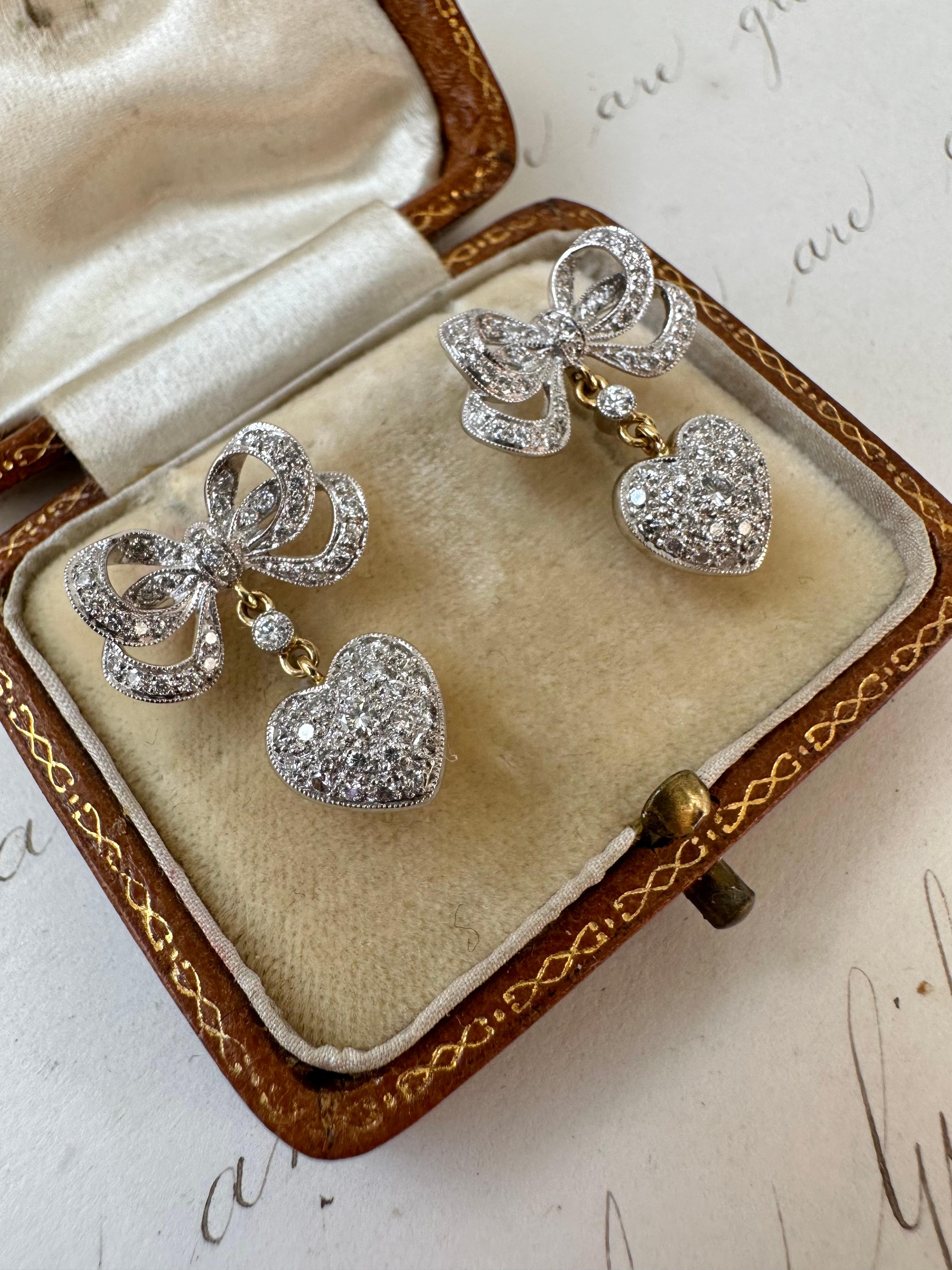 Sweet and symbolically beautiful, these contemporary crowned heart earrings feature a sparkling brilliant-cut diamond set heart, backed in rock crystal and crowned with a shimmering brilliant-cut diamond lined double bow. Crafted in 18K gold, these