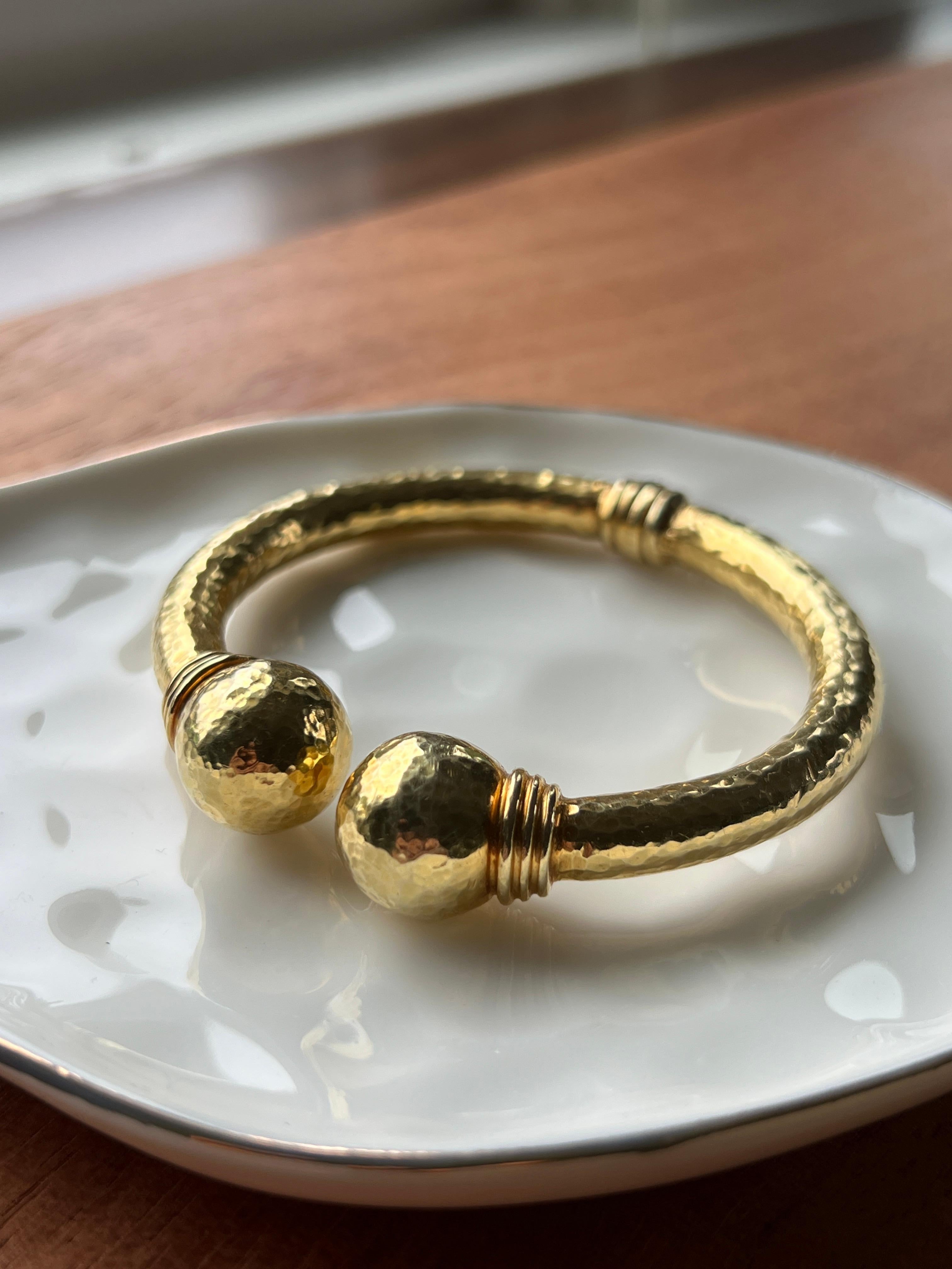Elegant and substantial, this contemporary bangle bracelet is crafted in solid 18 karat yellow gold and features a distinctive hammered finish that plays beautifully with light. The sleek hinged design is finished with polished ball terminals