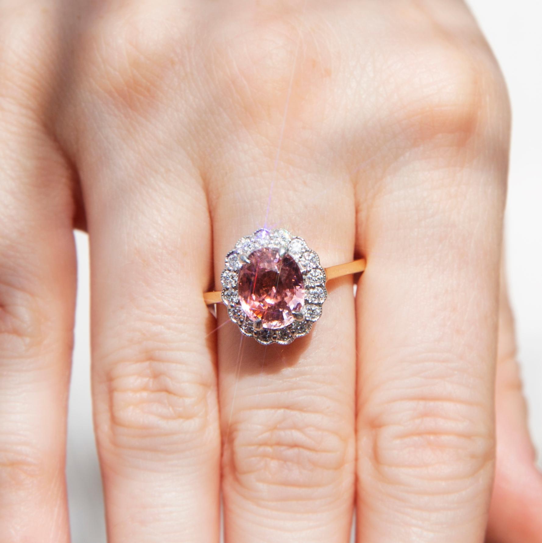 Lovingly crafted in 18 carat yellow and white gold is this alluring halo cluster ring featuring a stunning oval cut 2.41-carat bright peachy pink tourmaline and is set with a scalloped border of shimmering round brilliant cut diamonds. We have named