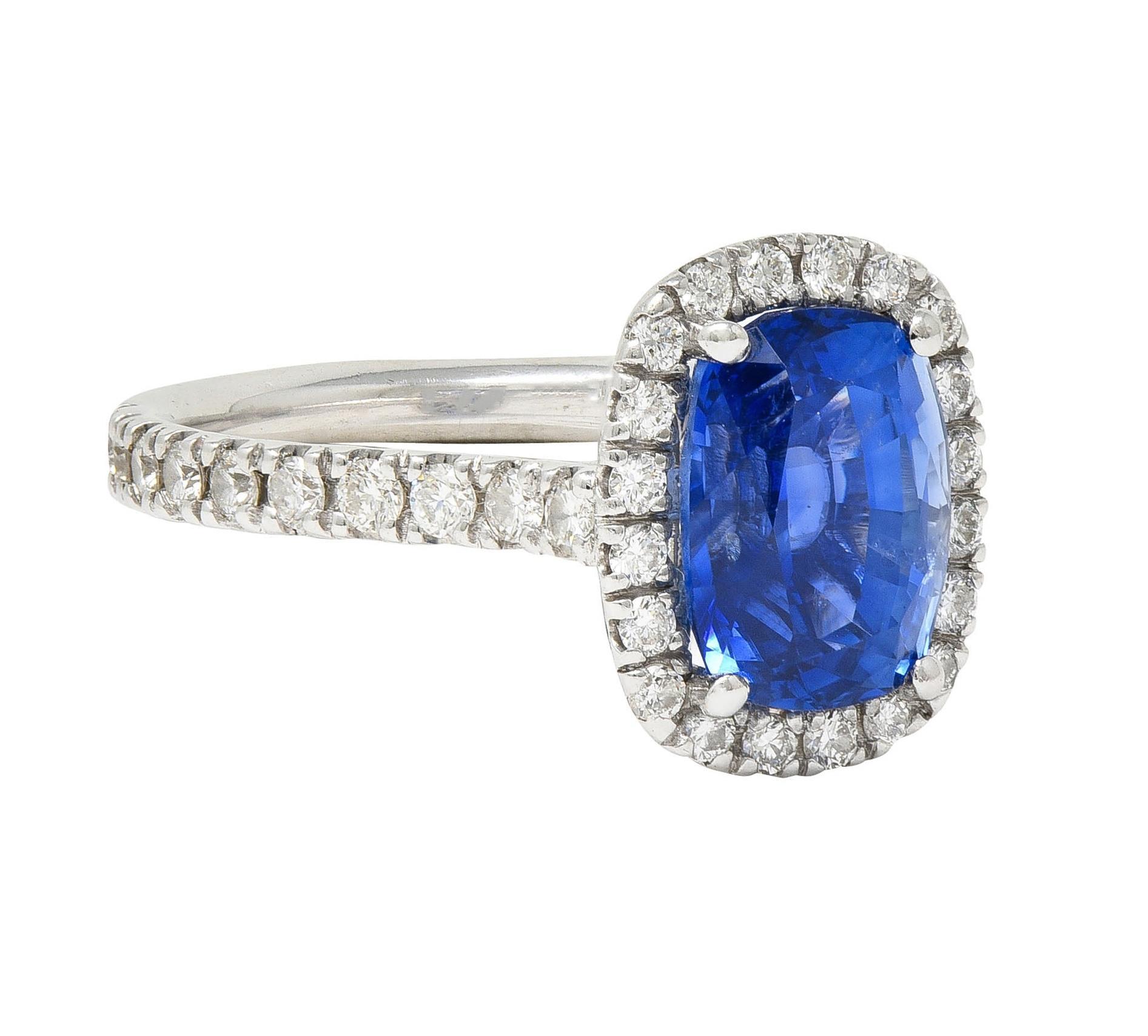Centering an elongated cushion cut sapphire weighing approximately 3.77 carat total 
Transparent medium vibrant blue in color - prong set in basket 
With a halo surround flanked by pierced cathedral shoulders 
Prong set with round brilliant cut
