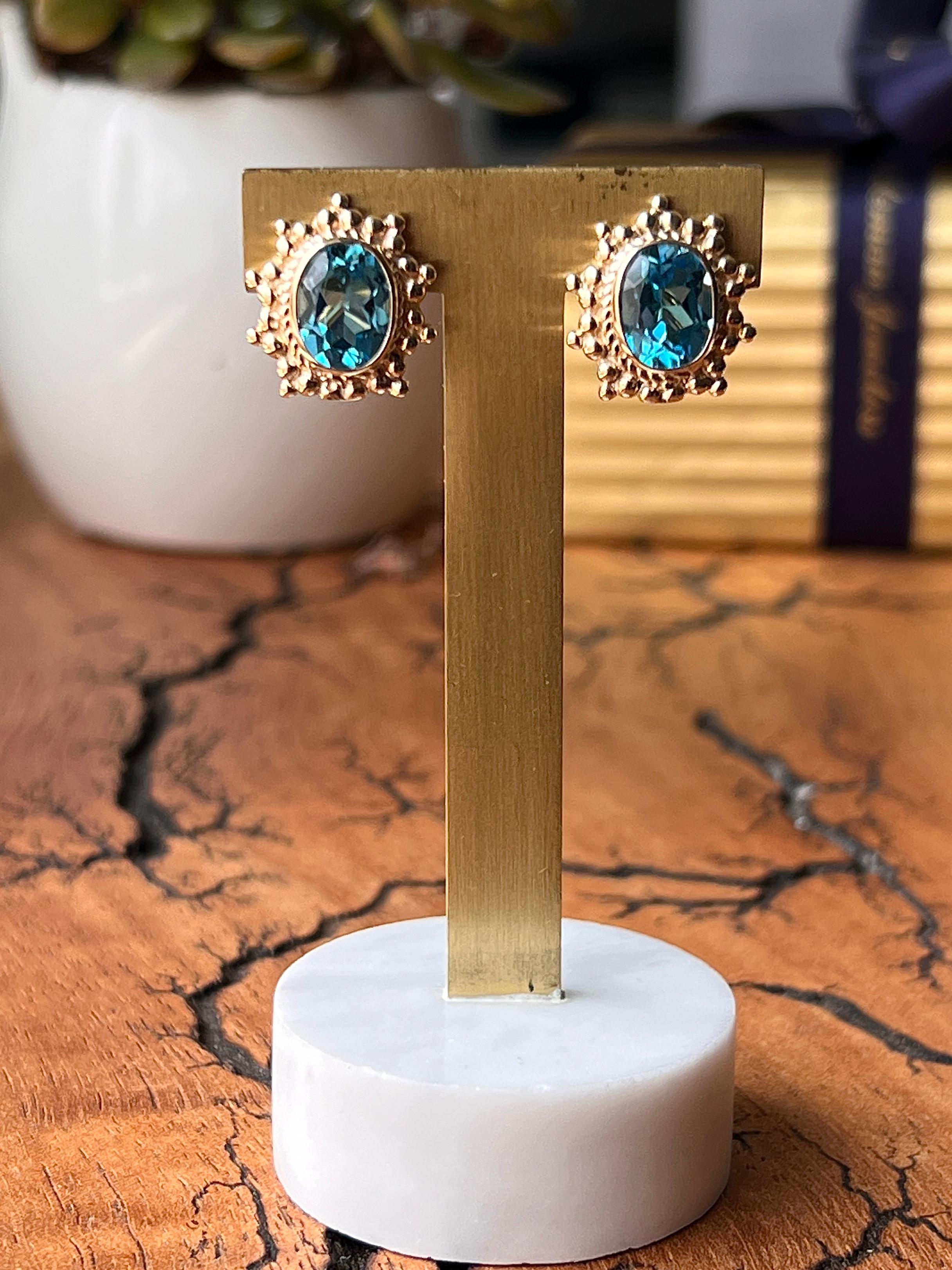 Un paio di orecchini contemporanei in oro giallo 14 carati, ciascuno con un topazio blu ovale che misura circa 9,8 x 7,4 x 5 mm, per un peso complessivo di circa 5,24 carati. Le gemme sono incorniciate in un bordo d'oro testurizzato e fissate con