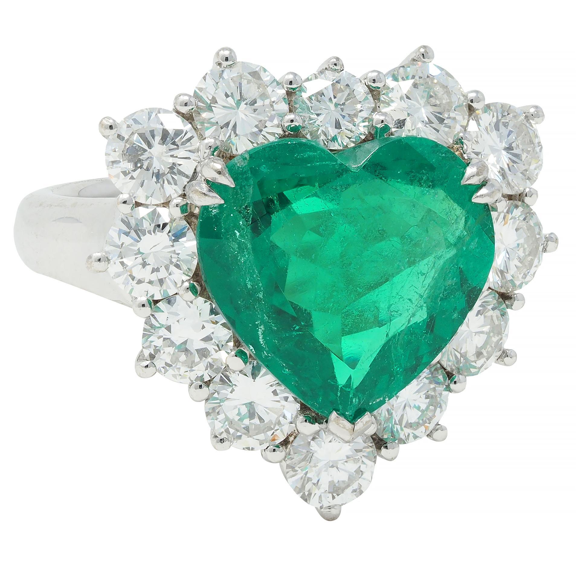 Centering a heart cut emerald weighing 4.62 carats - transparent medium green in color
Natural Colombian in origin with minor modern clarity enhancement 
Set with split talon prongs with a round brilliant cut diamond halo surround 
Weighing