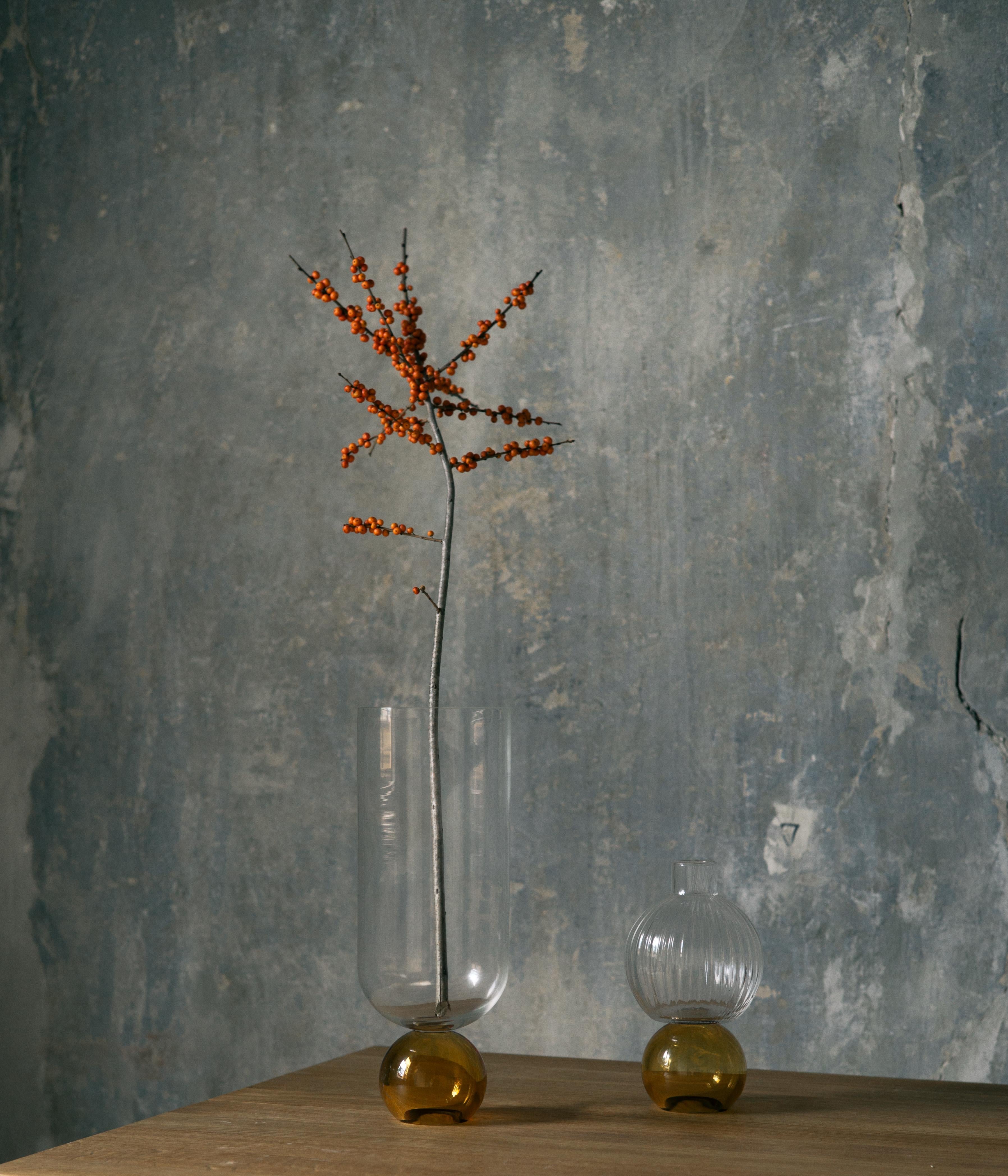 Diese bezaubernde, handgefertigte Vase aus feinem Glas ist ein Schaufenster des reinen Volumens. Ihre elegante Silhouette besteht aus einer kugelförmigen, grünen Basis, die den zylindrischen, klaren Körper trägt. Nach traditionellen Techniken aus