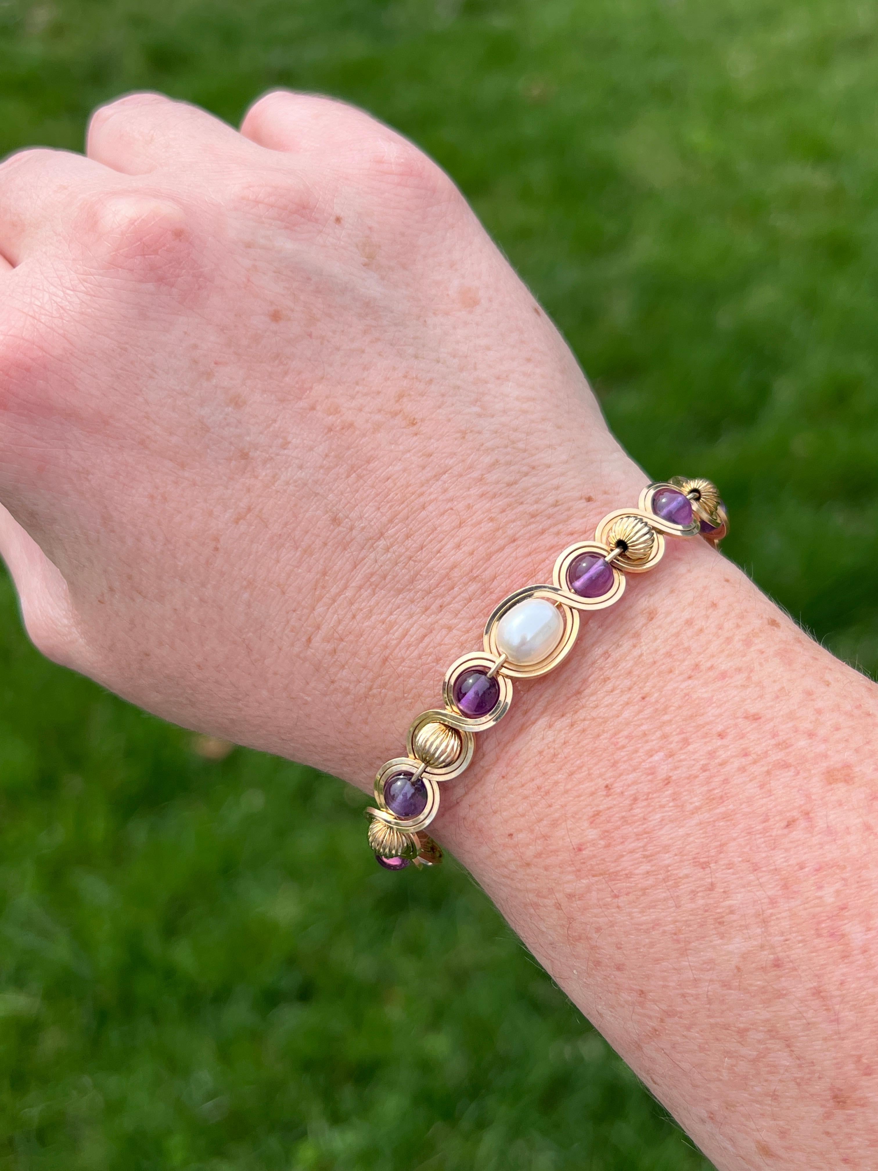 A graceful 14 karat yellow gold bracelet featuring six luminous 6mm round amethysts, a single 7.5mm pearl, and polished gold beads, all delicately beaded and wrapped with gold wire. The combination of gemstones and gold accents creates a rich