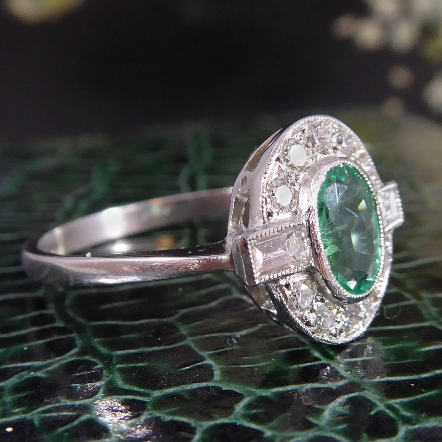 An emerald and diamond set cluster ring centring on a fully rub over set, oval shaped, mixed cut emerald estimated to weigh 0.70ct and measuring approx. 6.90mm x 5.20mm x 2.95mm deep, to a millegrain edged oval bezel collet.  A surround of 10 grain