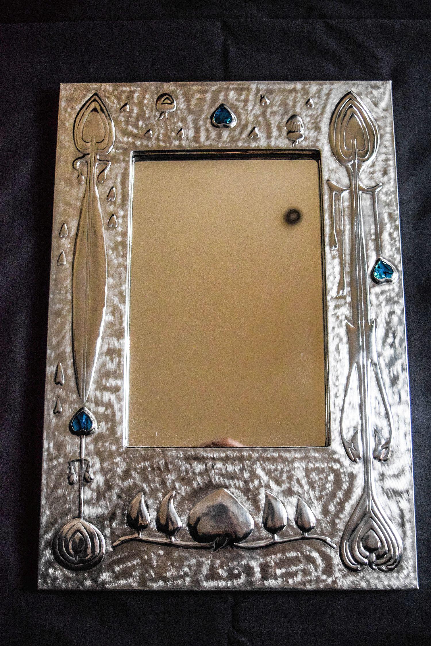 Contemporary Arts and Crafts Pewter Mirror with blue Cabuchons
A contemporary Arts and Crafts ornate pewter mirror with blue enamel cabuchons made by
M.Santos.Marked M.Santos on the edge and signed on the back.Also marked “HEART and 
Spoons.Abalone
