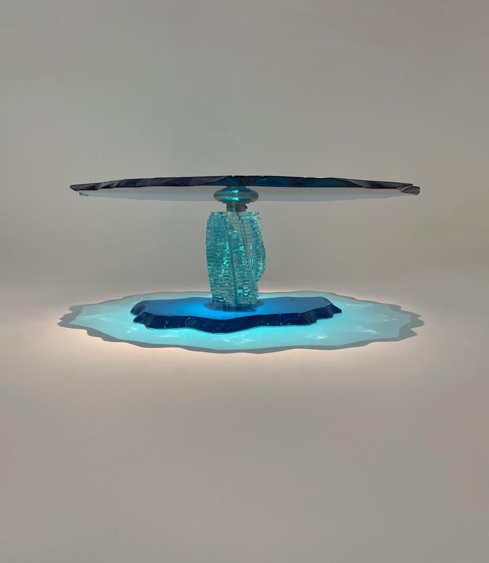 Danny Lane Studio poured turquoise-aquamarine deeply coloured glass (table top and base), the edges hammer fractured and finished by hand polishing.

The post-tensioned stacked and carved low iron glass column joins top and base with stainless steel