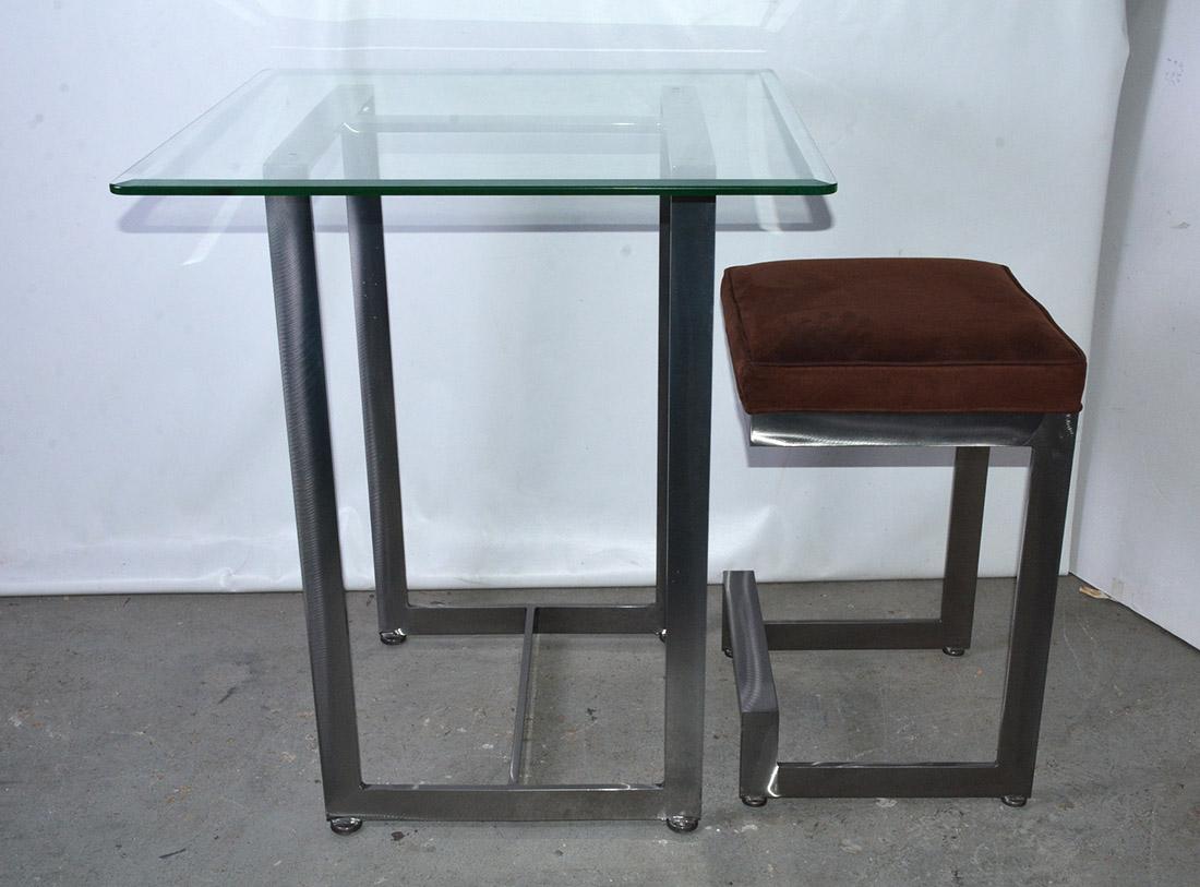 The contemporary Mid-Century Modern bar table is made of a welded stainless steel frame with a beveled glass top. The matching bar stool consists of a welded stainless steel frame topped by a brown faux suede cushion. Both pieces have padded