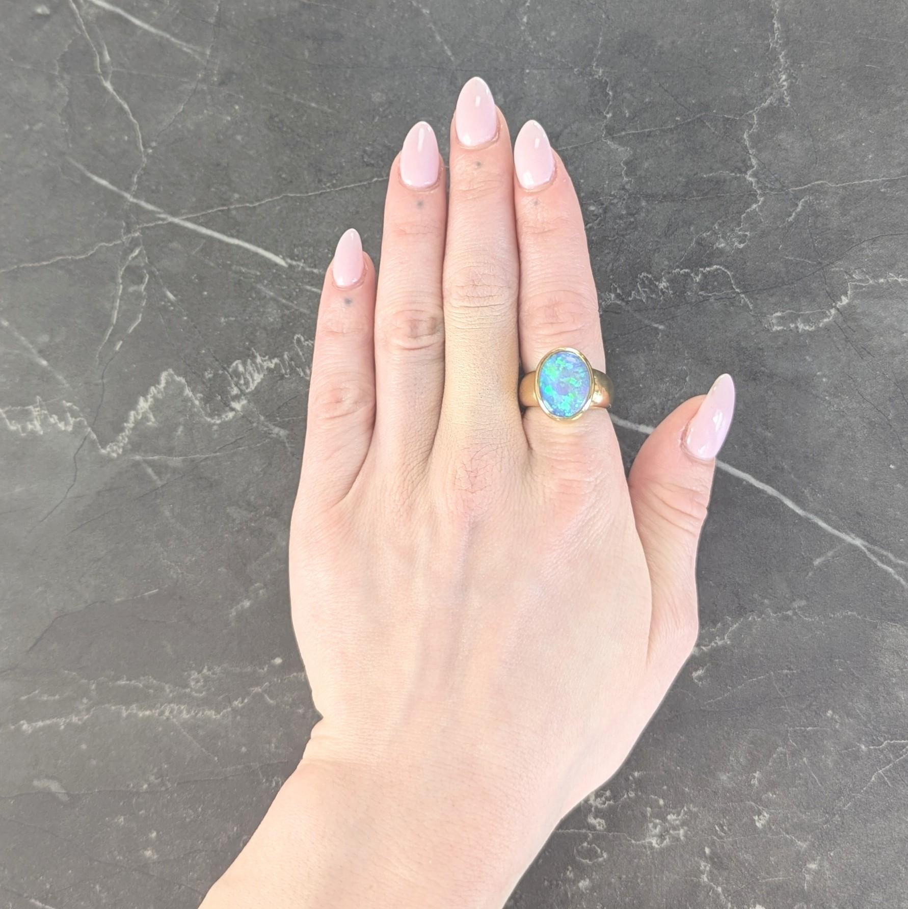 Centering an oval shaped opal cabochon measuring 11.5 x 15.0 mm
Transparent black in body color with strong blue/green/violet flash
Bezel set and flanked by puffy rounded shoulders
With a high polish finish
Stamped for 18 karat gold
With maker's