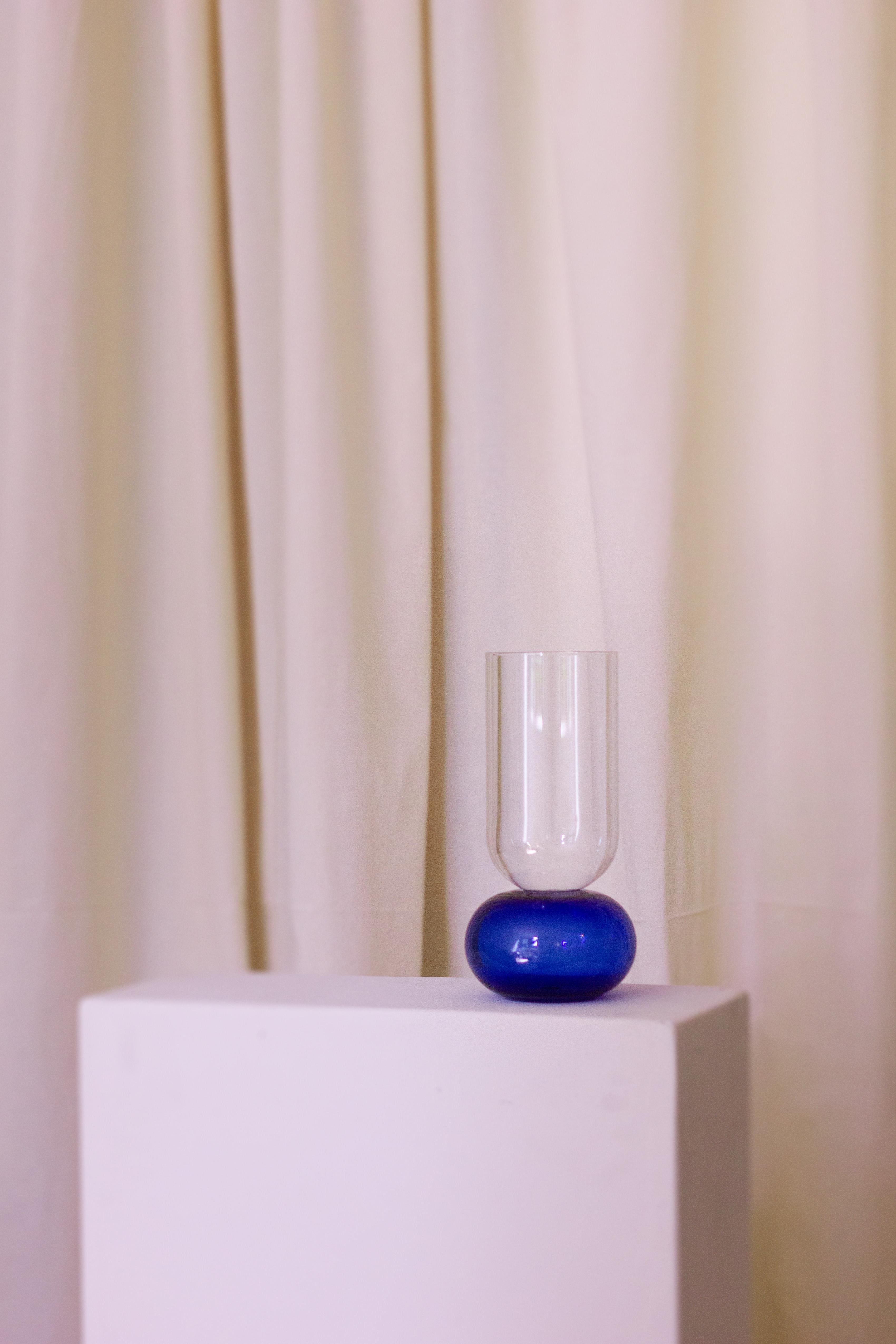 Diese bezaubernde, handgefertigte Vase aus feinem Kristall ist ein Schaufenster der reinen Volumen. Ihre elegante Silhouette besteht aus einem kugelförmigen, blauen Sockel, der den zylindrischen, klaren Körper trägt. Nach traditionellen Techniken