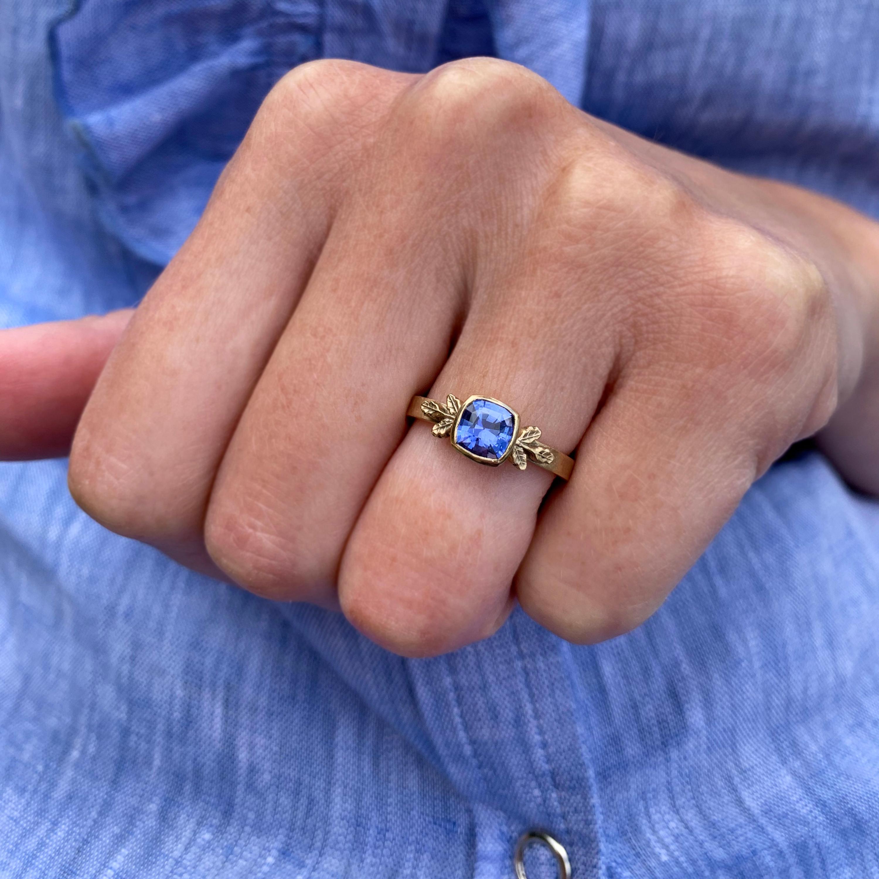 Cornflower blue sapphire set in yellow gold with signature leaf detail – simply exquisite ring. 
A stunning cushion cut sapphire is set in a full bezel and surrounded with Beth’s signature leaf details on either side. The incredible purple blue hue