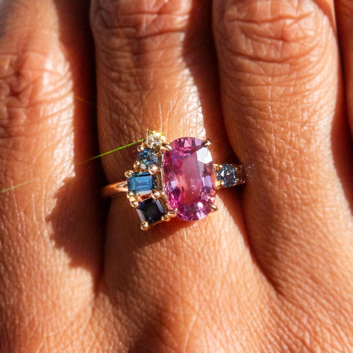 Thoughtfully crafted with love and intention, this 18 carat gold is a gorgeous contemporary adornment. At the centre is a lovely oval cut purple spinel with an alluring asymmetrical fanned cluster of emerald cut natural blue and teal sapphires and