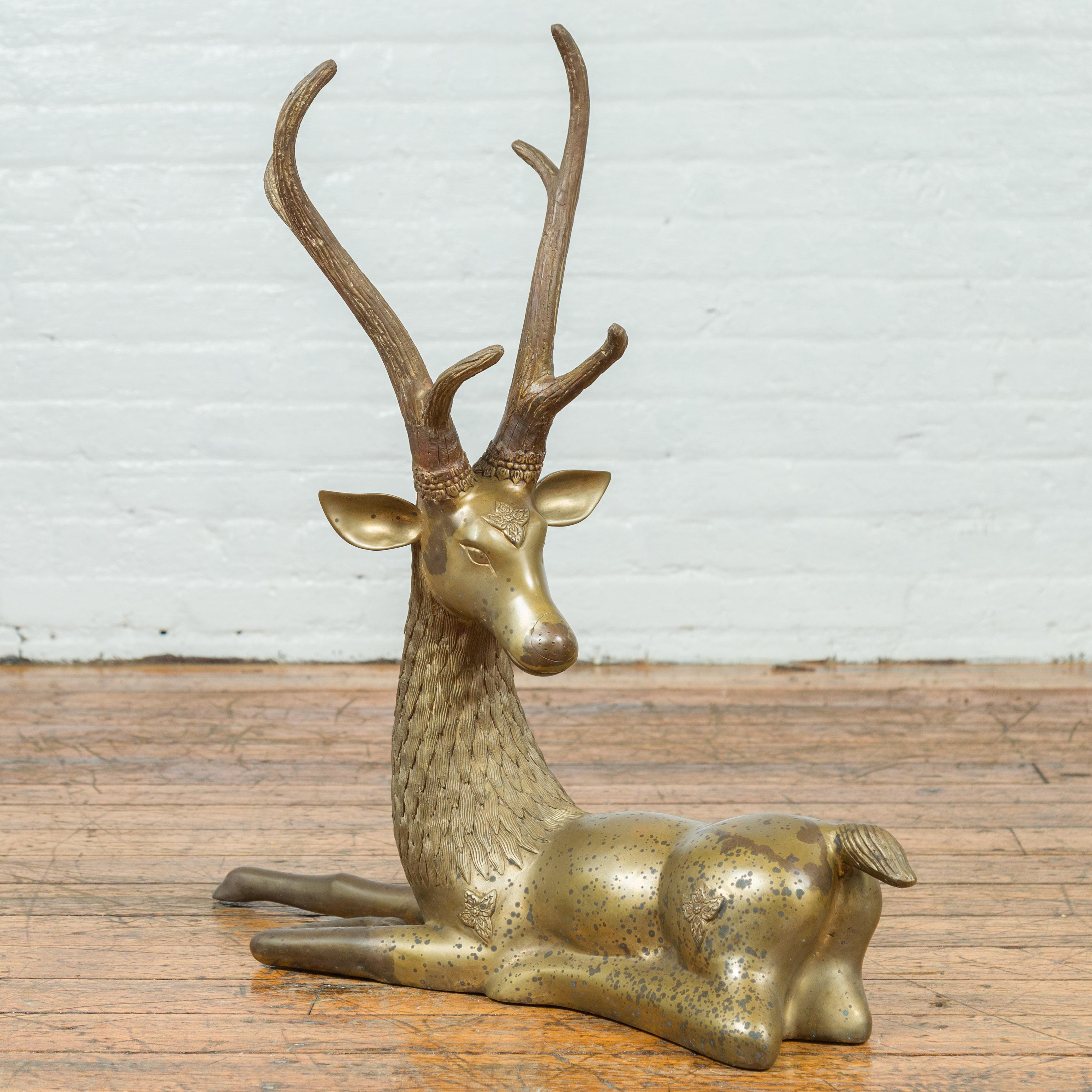 A contemporary bronze reclining deer statue with golden patina, large antlers and floral ornaments. Created with the traditional technique of the lost-wax (à la cire perdue) that allows a great precision and finesse in the details, this statue