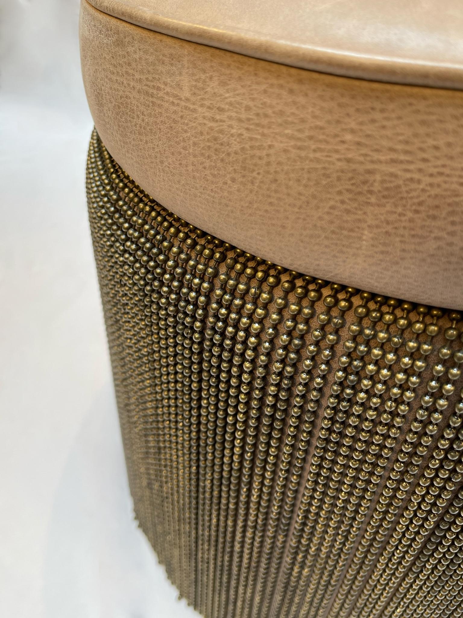 A contemporary ottoman stool, handcrafted in London in 2010 by Spinna Design. The generously proportioned circular form is upholstered in a warm-toned brown cowhide leather, edged in matching piped trim. Around the body, a dense bronze metal ball