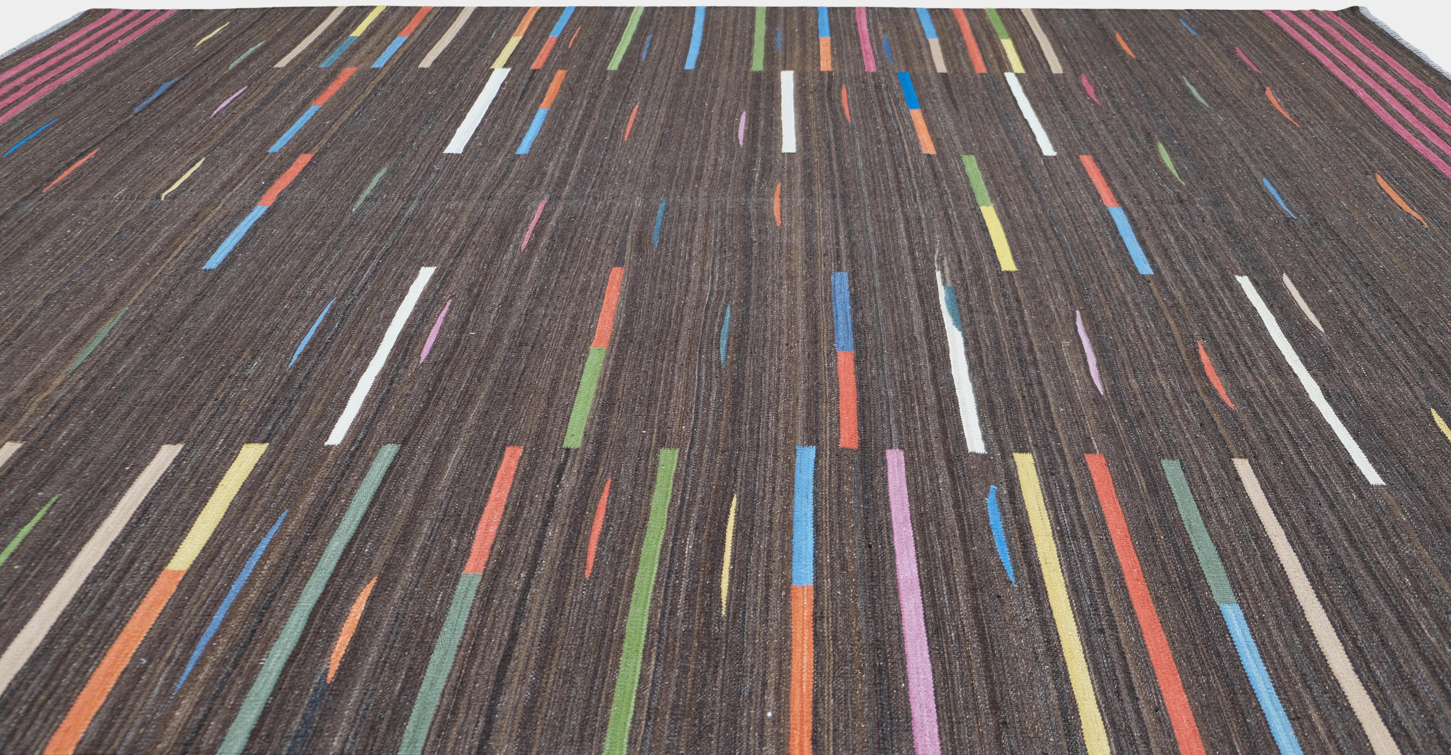 Eleva tu espacio con esta alfombra de lana tejida a mano en Pakistán, con un rico fondo marrón acentuado por vibrantes motivos lineales multicolores en tonos naranja, azul, verde, amarillo, blanco y rosa. Su diseño elegante y minimalista, aunque