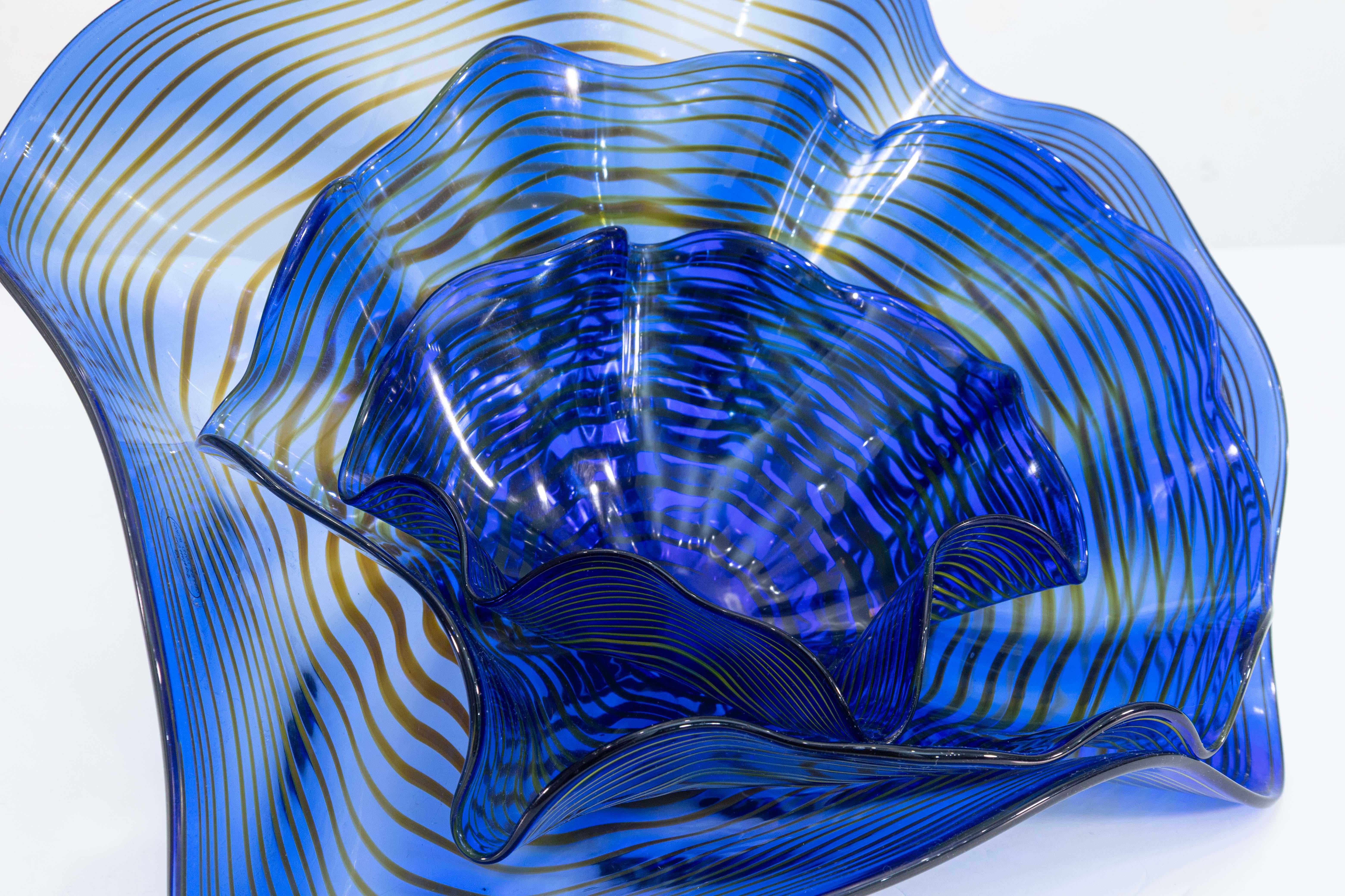 A Viz Glass Inc Chihuly style 3pc glass table sculpture. The sculpture is made from a lovely hand blow, blue with striped layers of yellows. The abstract shapes of each of the glass sculptures sit inside each other to form layers of waving glass.