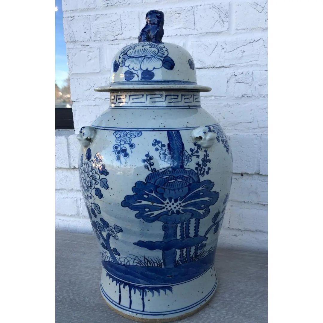 This unique, handcrafted temple jar is painted with a blue and white floral tree pattern. The jar is shaped by bold curves and is topped with a lid, adorned with blue lion figurine. Traditional Chinese markings line the top rim and the edge of the
