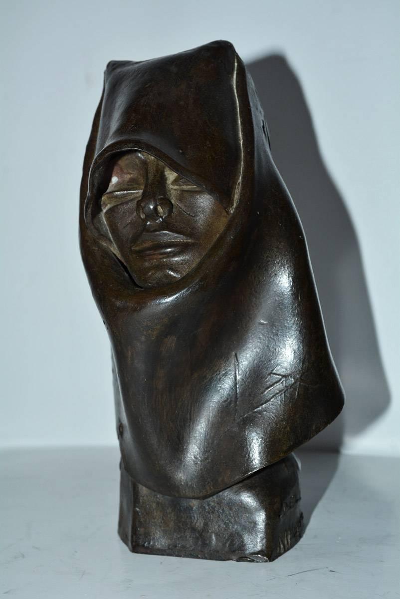 The contemporary sculpture in clay represents a female head swathed in fabric for a hooded effect. The figure is hollow. The head sits on a stand with the engraved name of Eli Nuoro.