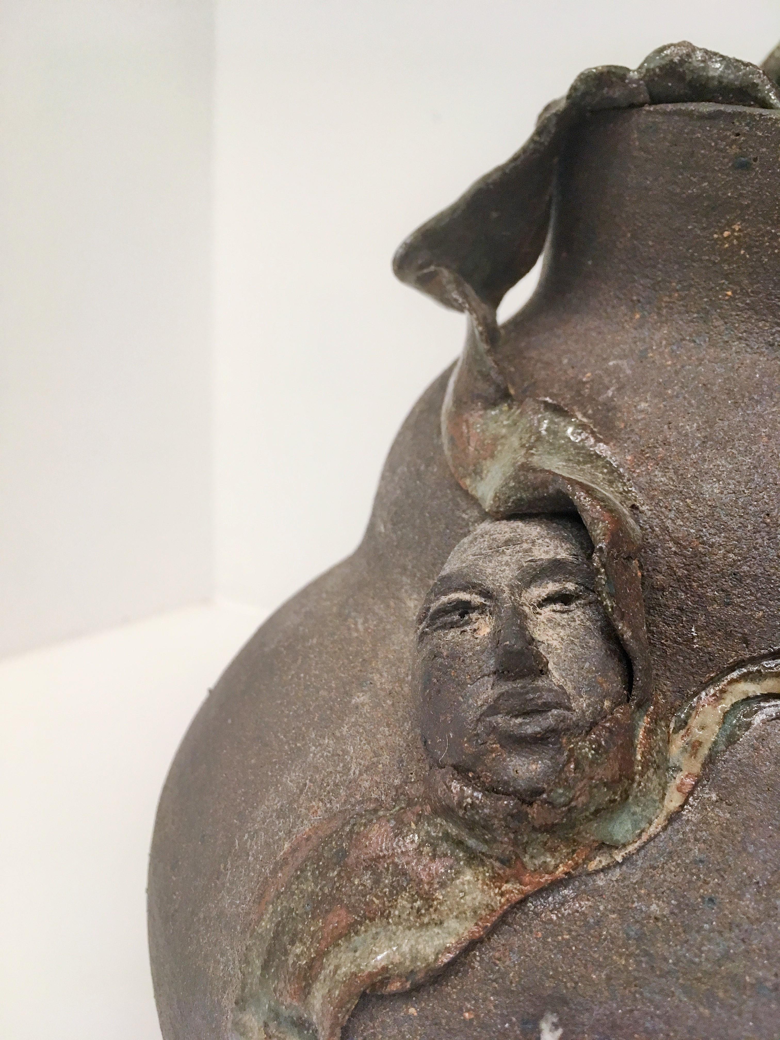 Contemporary clay vessel, with brownish-green glaze. A figure enveloped in clay takes center stage. Signed.