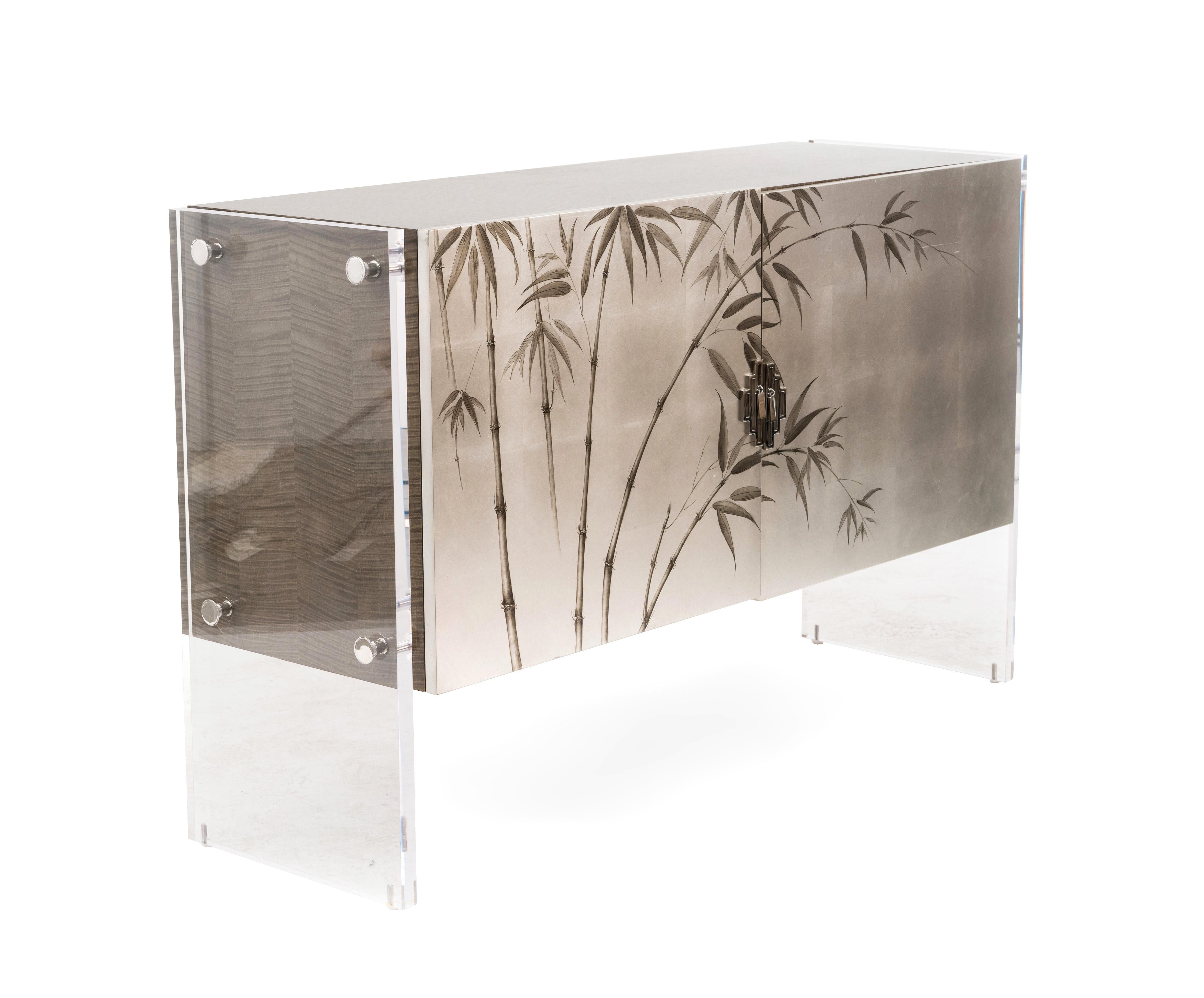 Contemporary silver/gray painted commode with bamboo floral design on the front doors and a gray wood veneer finished top. Sides supported on solid Lucite panels with silver metal detail. (Theodore Alexander).


Theodore Alexander is one of the