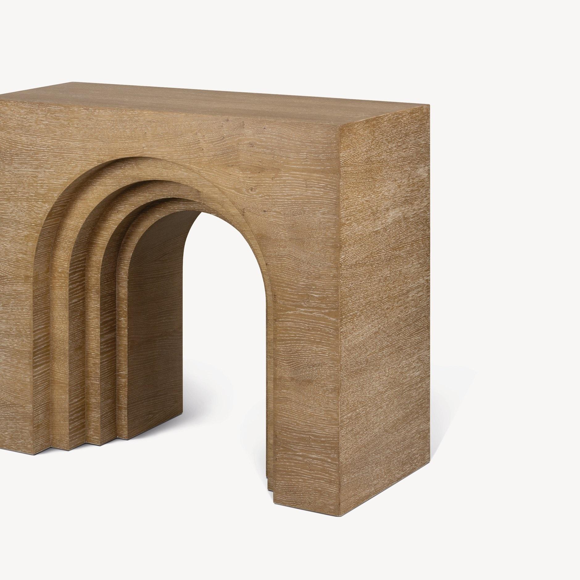 This console is made of limed oak wood and has a distinctive, arched or bridge-like shape. The console table has a smooth, polished finish and sits on two slender legs. It is characterized by its organic shapes and use of natural materials. The