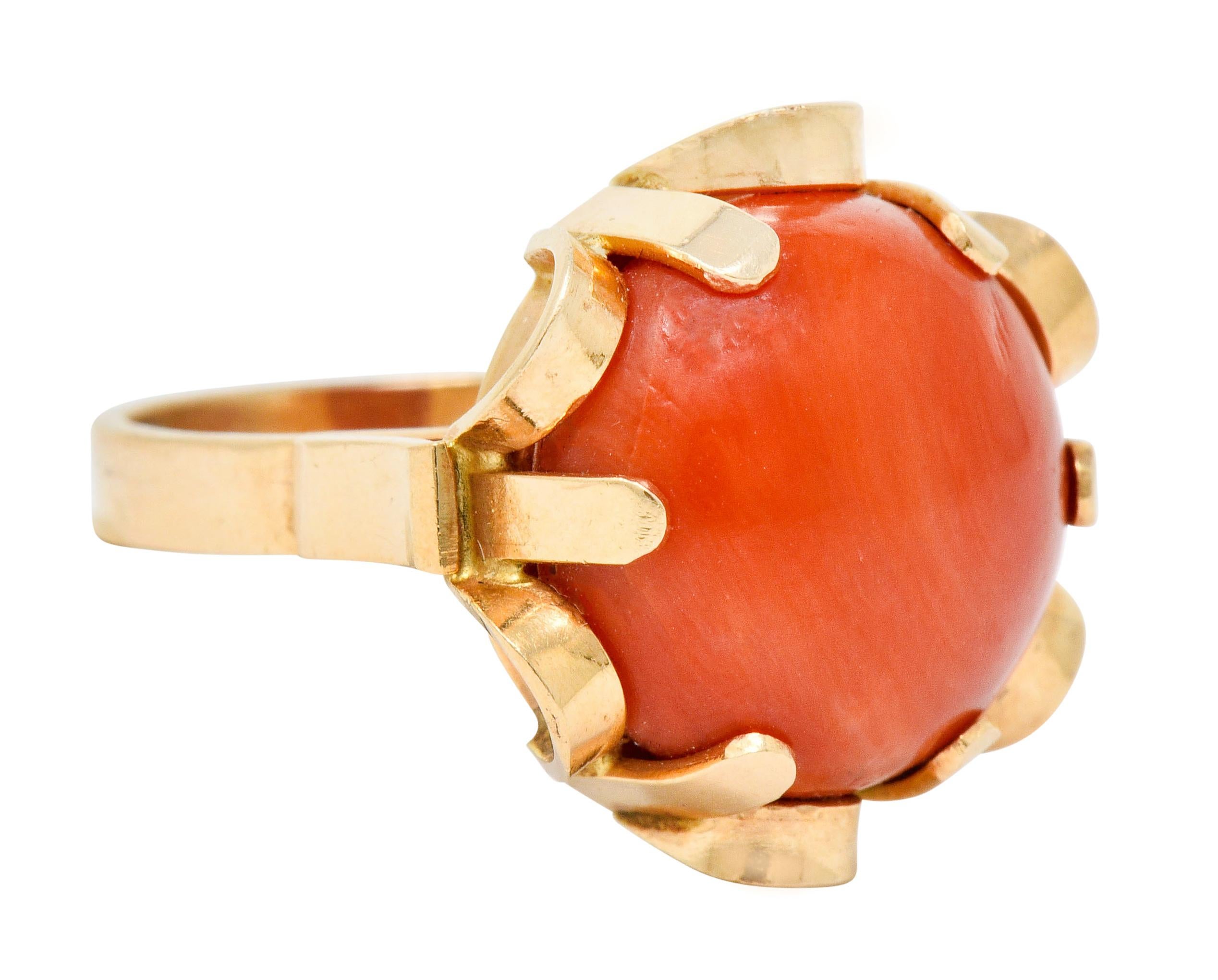 Centering a round coral cabochon, robust reddish-orange in color; with good surface quality and luster

Prong set and surrounded by a stylized and dynamically looped floral motif

Tested as 14 karat gold

With Polish assay marks, possibly for
