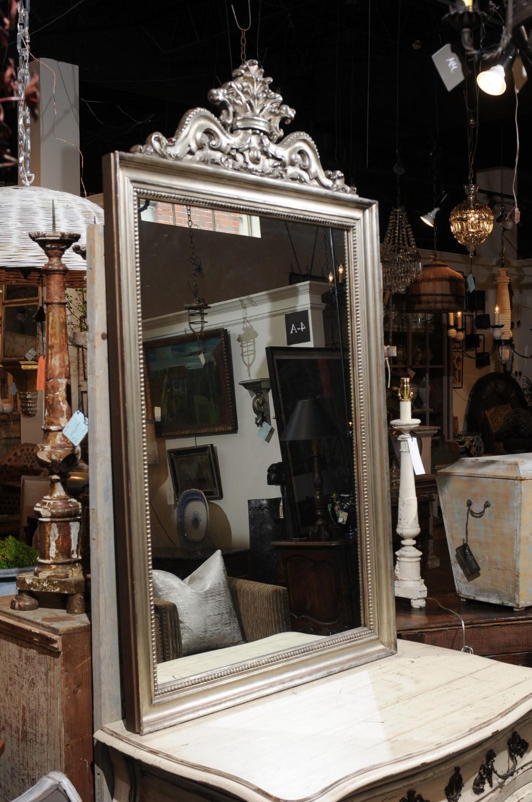 A contemporary Rococo style crested mirror framed in antiqued silver, with Rocailles motifs. Capturing our attention with its skillfully carved crest and silver finish, this mirror features a rectangular molded frame accented with delicate beads on