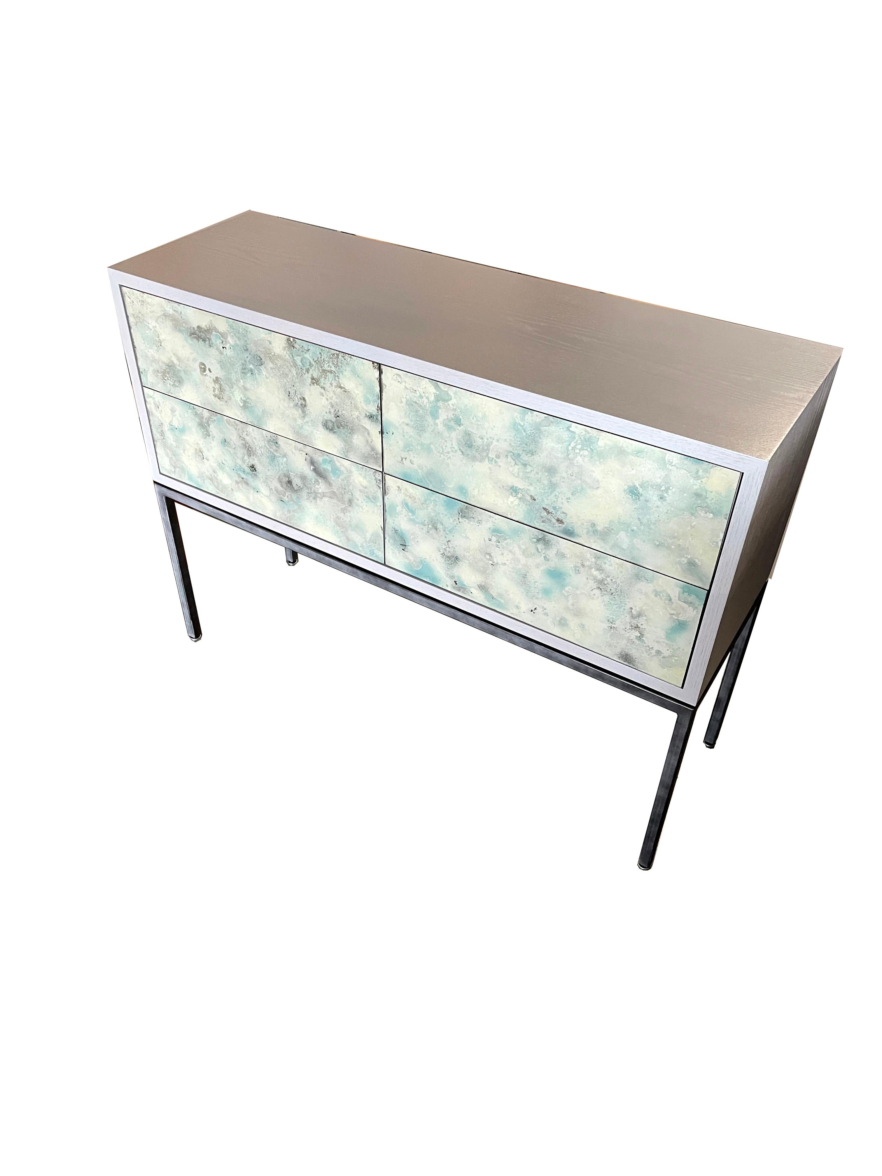 The églomisé chest of drawer / Console by Ercole Home has a 4-drawer front with a morning gray finish on oak.
 Hand painted églomisé glass panels are on the surface of each drawer in our custom made celadon dust finish. Drawers with self-closing