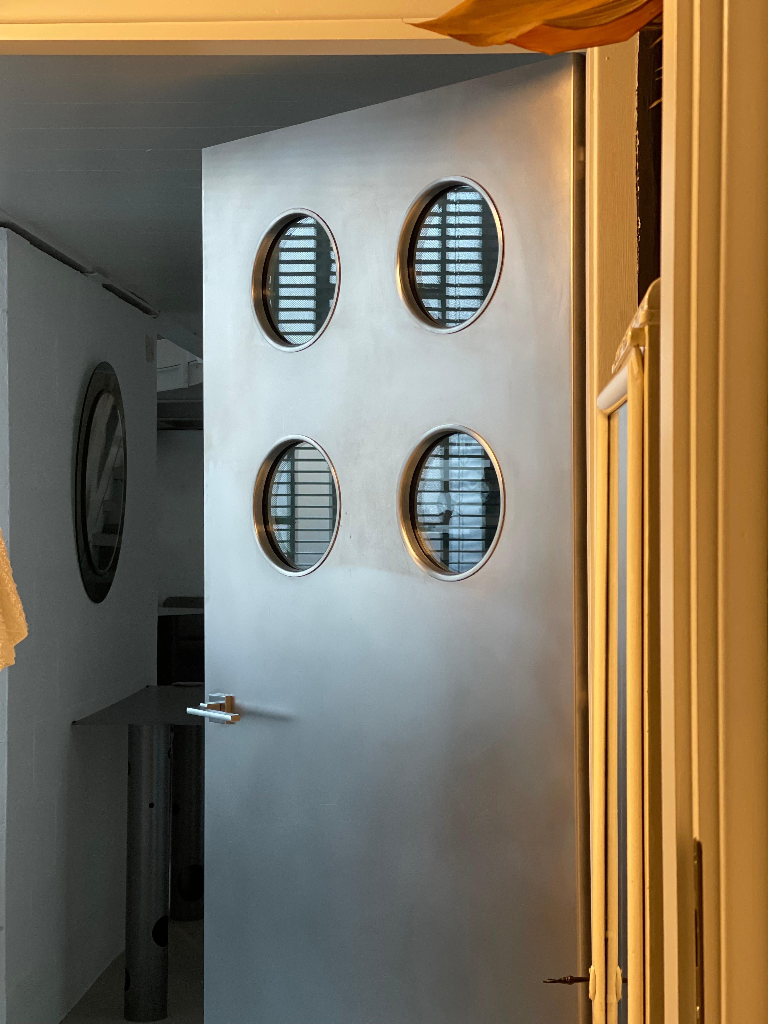 Contemporary custom made stainless steel metal door with round portholes, designed by Spinzi and made in Italy.

Inspired by the works of Jean Prouvé, this door is built around an existing regular wood door, with the addition of round porthole