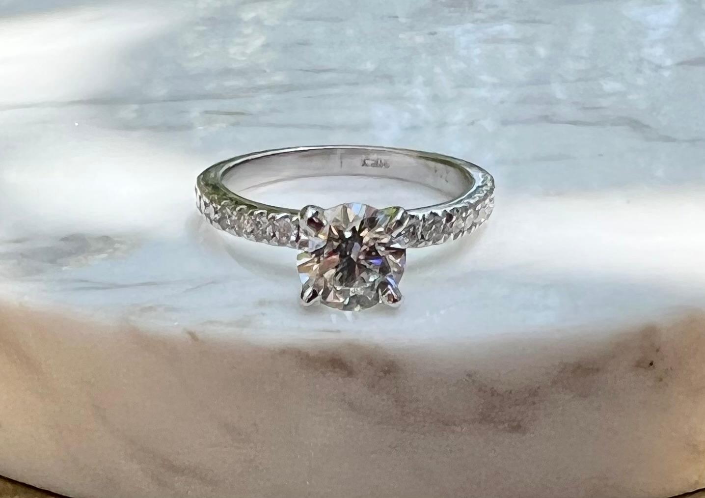 Un anello di fidanzamento in oro bianco 14 carati con un diamante rotondo taglio brillante da 1,00 carati con colore H e purezza SI2.  La pietra centrale è incastonata in una montatura con 0,27 carati di diamanti taglio brillante con colore G/H e