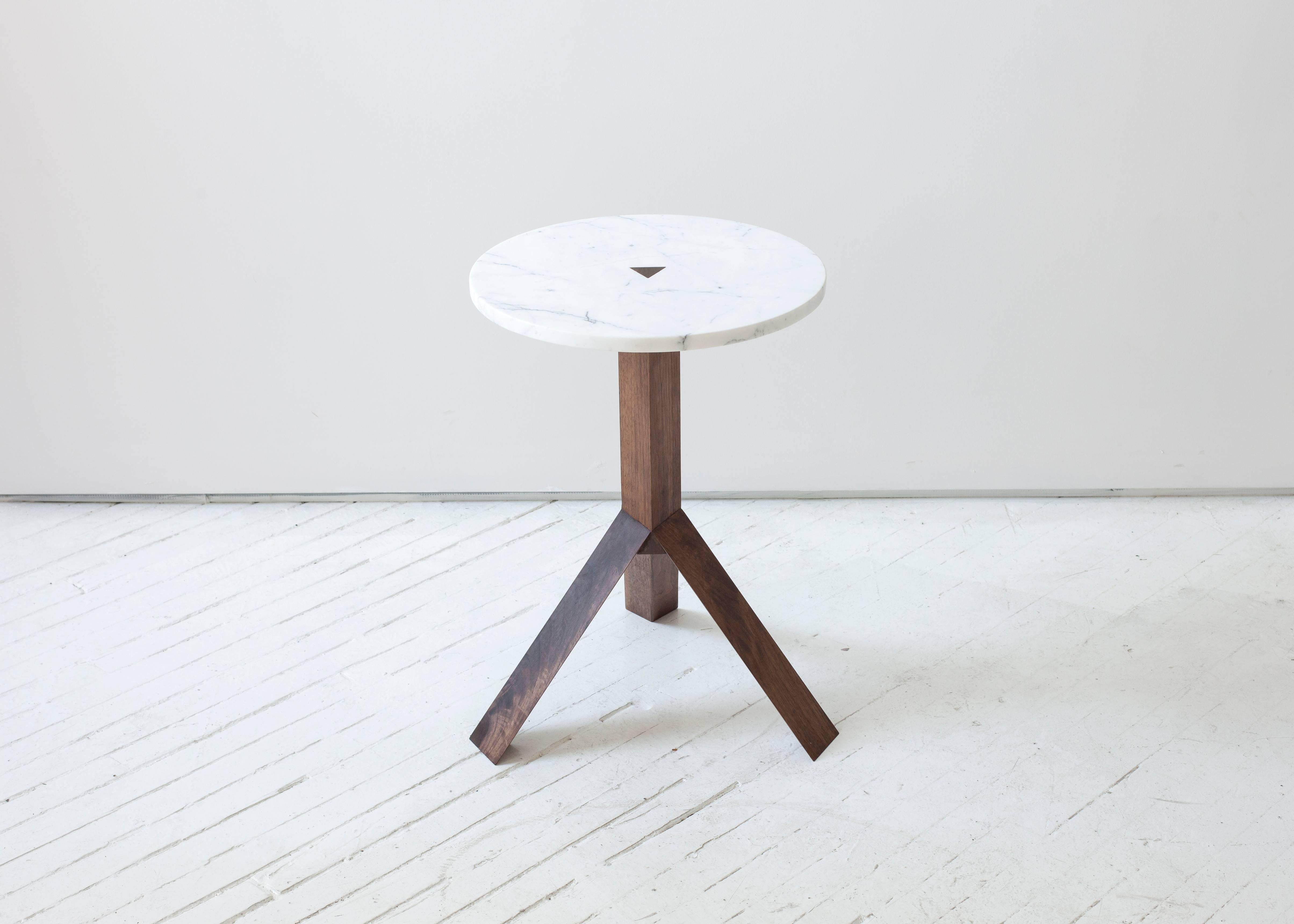 Three angular wood legs split off from a triangular centre post to elevate the small geometric stone tops of this side table. A triangular wood tenon pierces the centre of the table top, communicating the construction and creating a subtle, flush