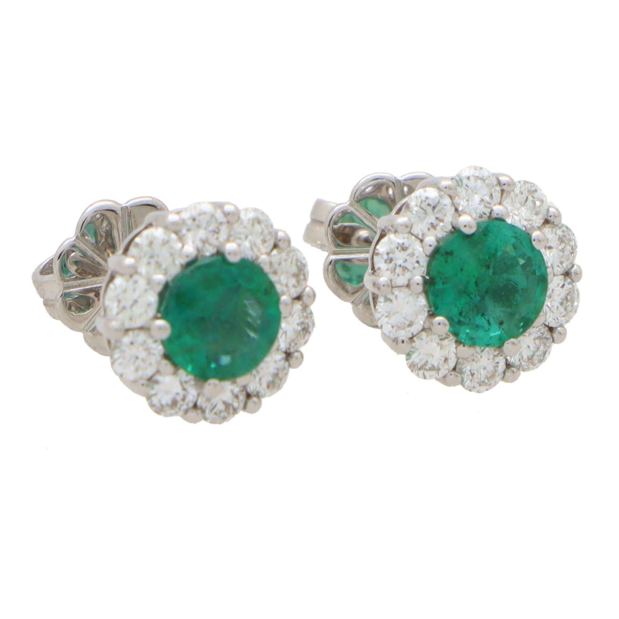 An elegant pair of emerald and diamond cluster stud earrings set in 18k white gold.

Each earring depicts a floral cluster motif centrally set with a vibrant emerald and surrounded by 10 round brilliant cut diamonds. The earrings are secured to