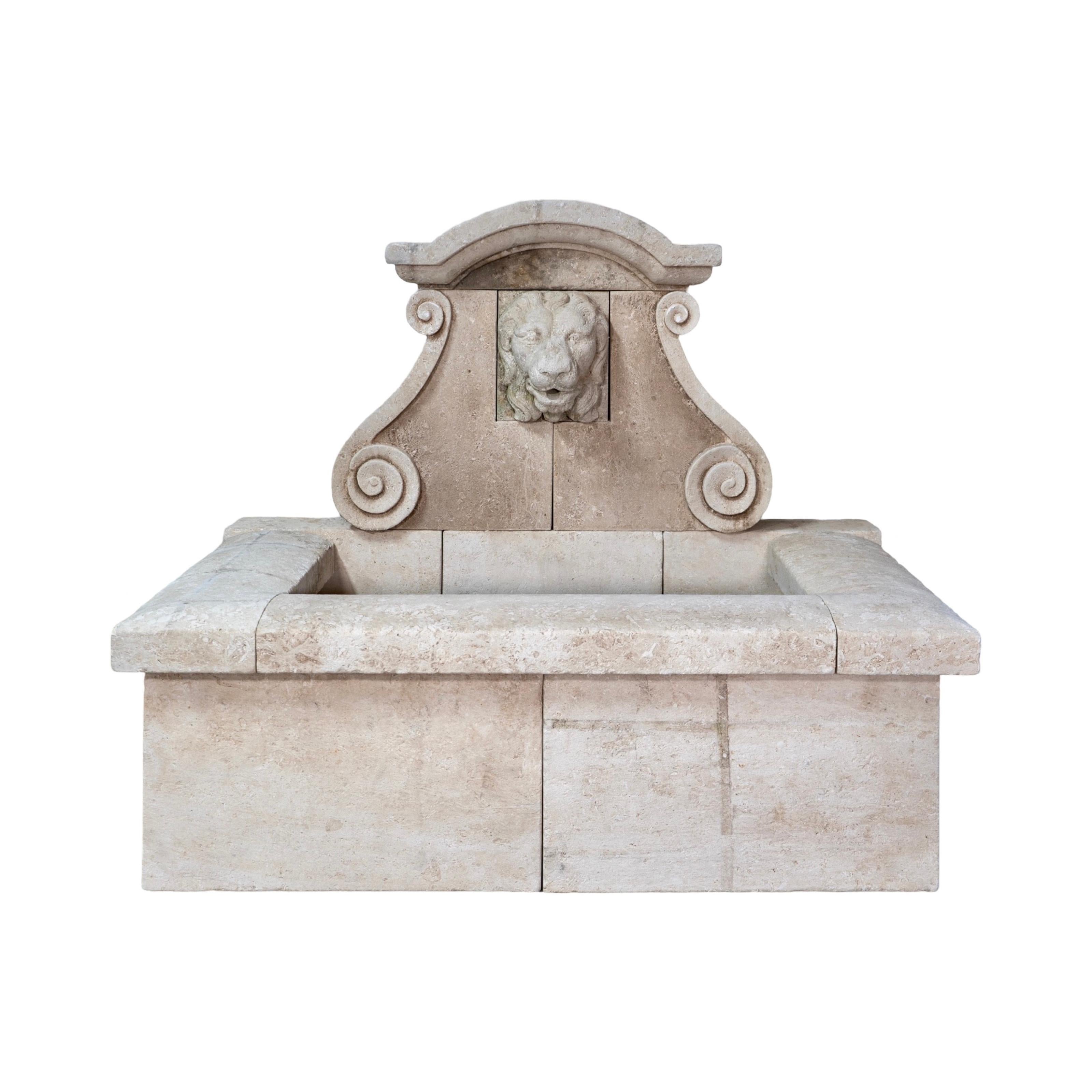 Une fontaine murale en pierre calcaire sculptée à la main qui présente la tête d'un lion sculptée dans la pierre calcaire comme sortie d'eau. Cette pièce est complétée par des volutes de chaque côté de la plaque arrière et par une couronne